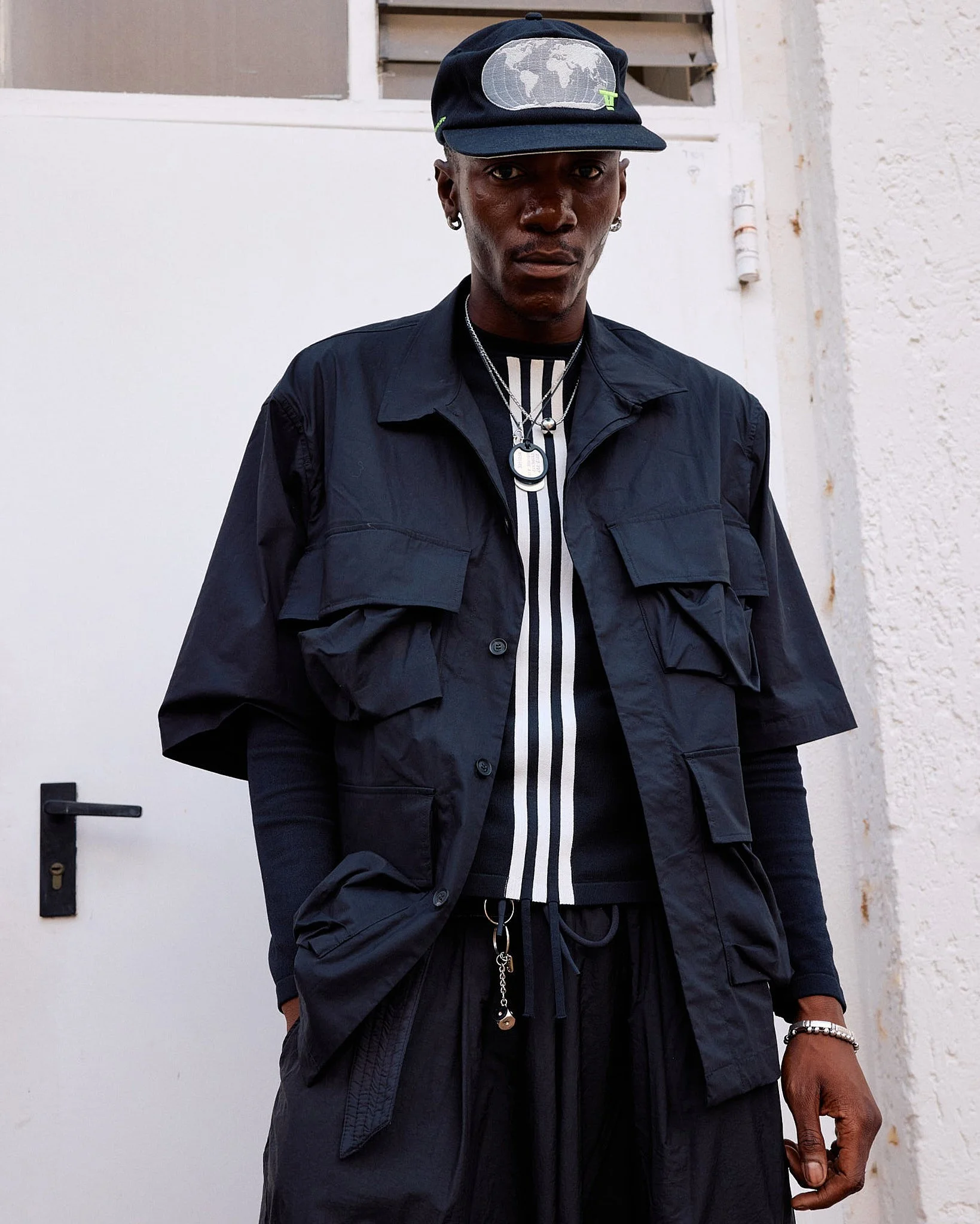 Menswear guest layering an oversized navy utility jacket with cargo flap pockets over a white-striped knit, drawstring trousers and a world-map cap, Berlin Fashion Week SS27