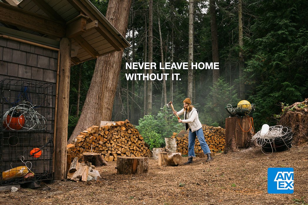 Advertising photographer London campaign image for speculative American Express campaign image of blonde actress chopping wood on tree stump outside forest cabin with crab traps in foreground and stacks of firewood.