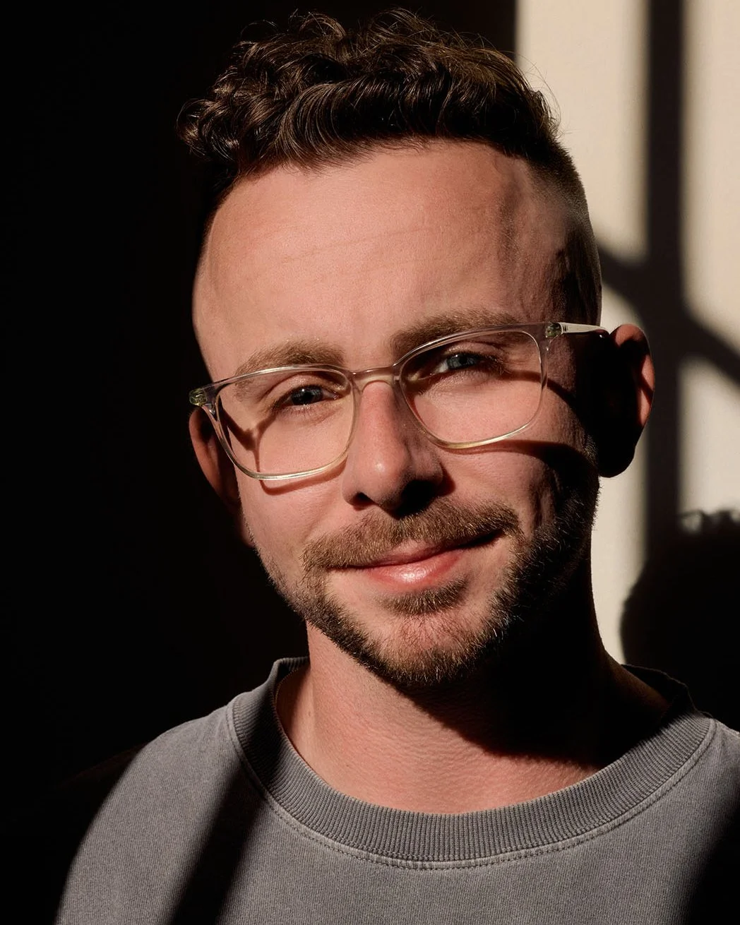 Lifestyle and street photographer London Ian Kobylanski portrait in direct sun window light with clear glasses, blue eyes, curly hair and grey shirt with confident expression looking at camera
