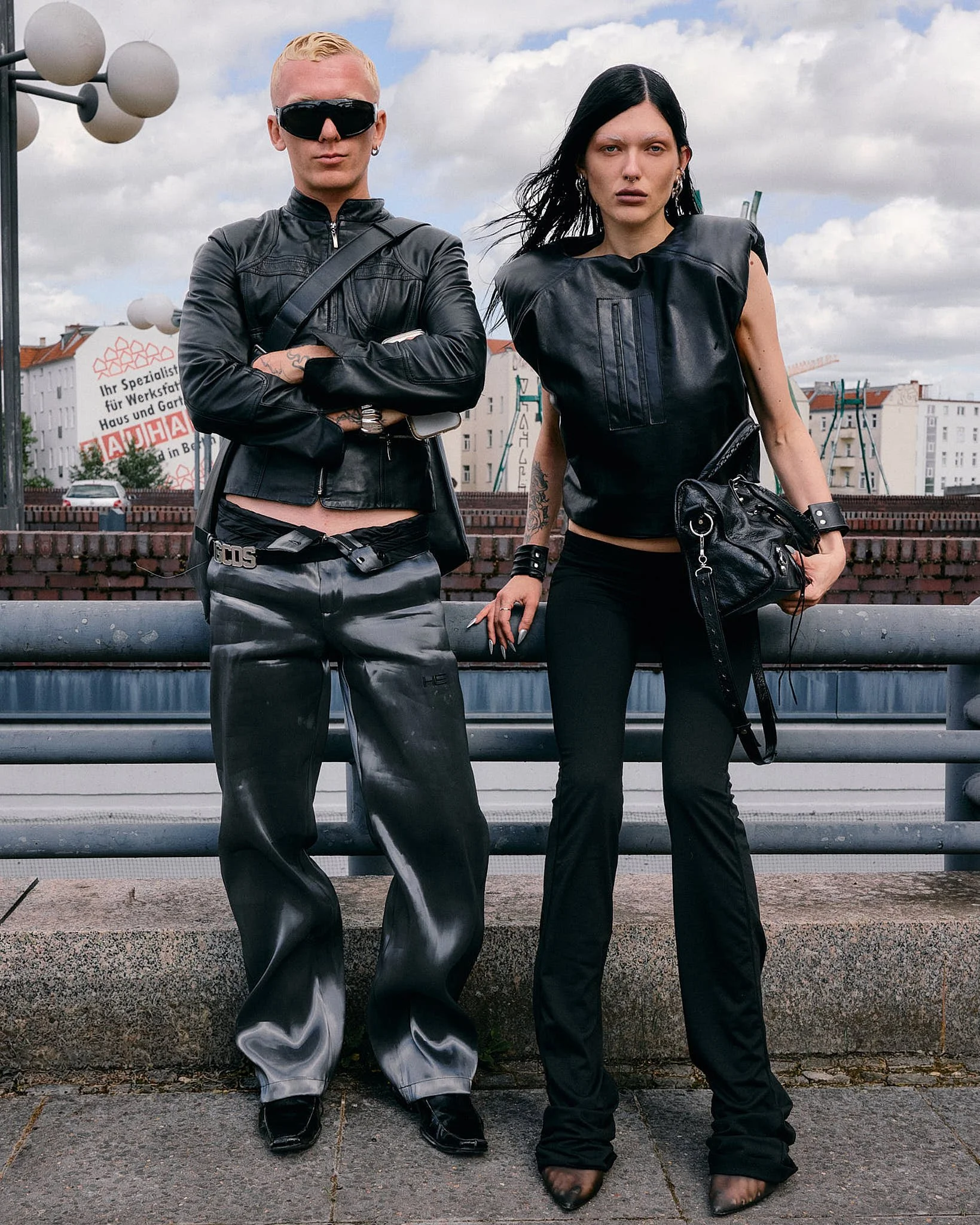 Two guests in head-to-toe black leather — cropped moto jacket over low-slung satin trousers, and a padded-shoulder leather top with a distressed motorcycle bag — on a Berlin bridge, Berlin Fashion Week SS27