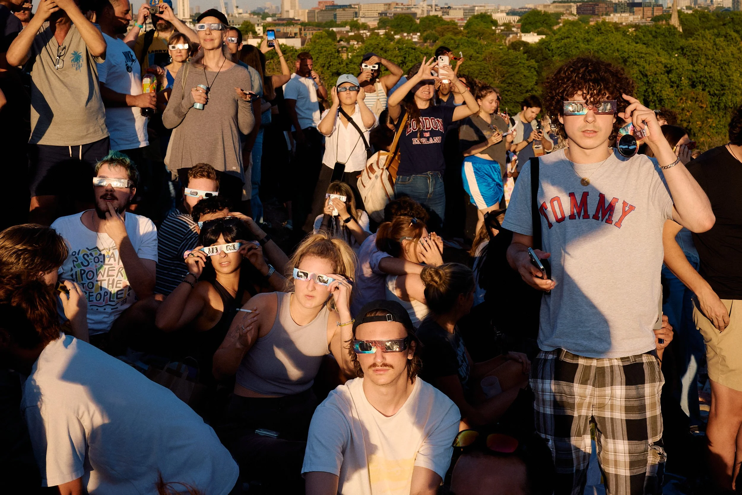 Crowd on Primrose Hill looking up through eclipse glasses at the solar eclipse, a young man in a Tommy T-shirt at the centre