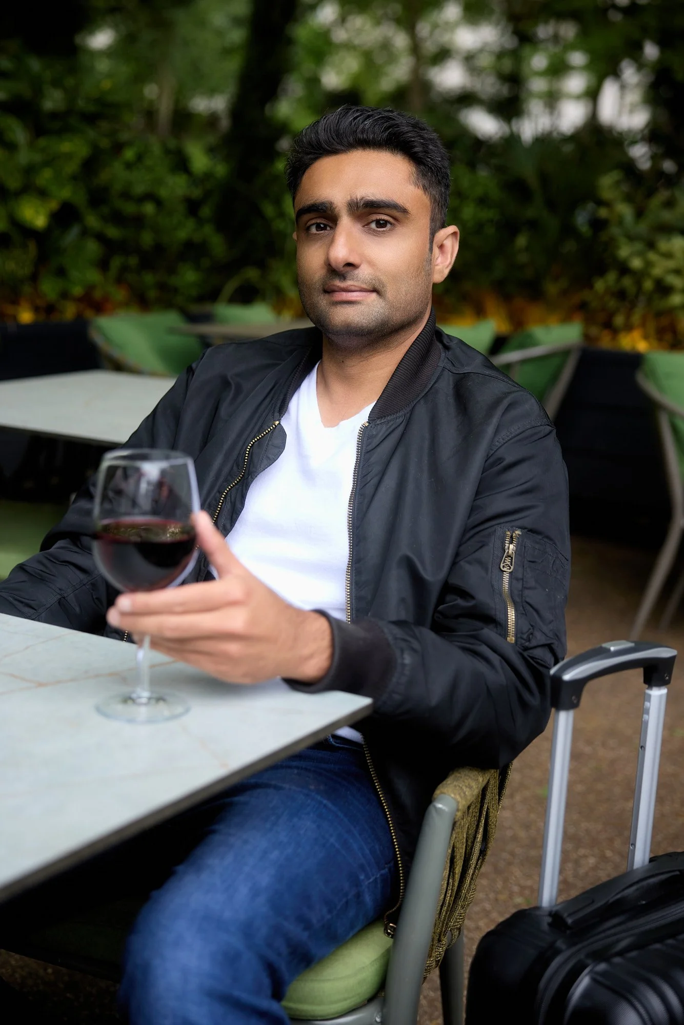 Man seated with glass of wine at restaurant with suitcase next to him as prop, as if he's waiting for a layover flight