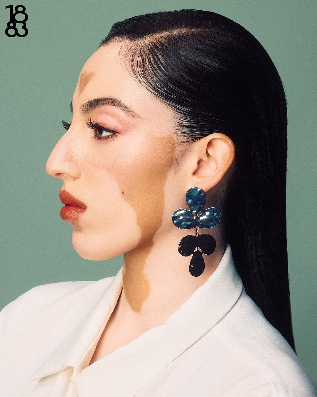 Commercial lifestyle photographer in London captures editorial fashion photography of female model in studio with vitiligo in studio with green painted wall, silver earring, white top looking away from camera close up portrait