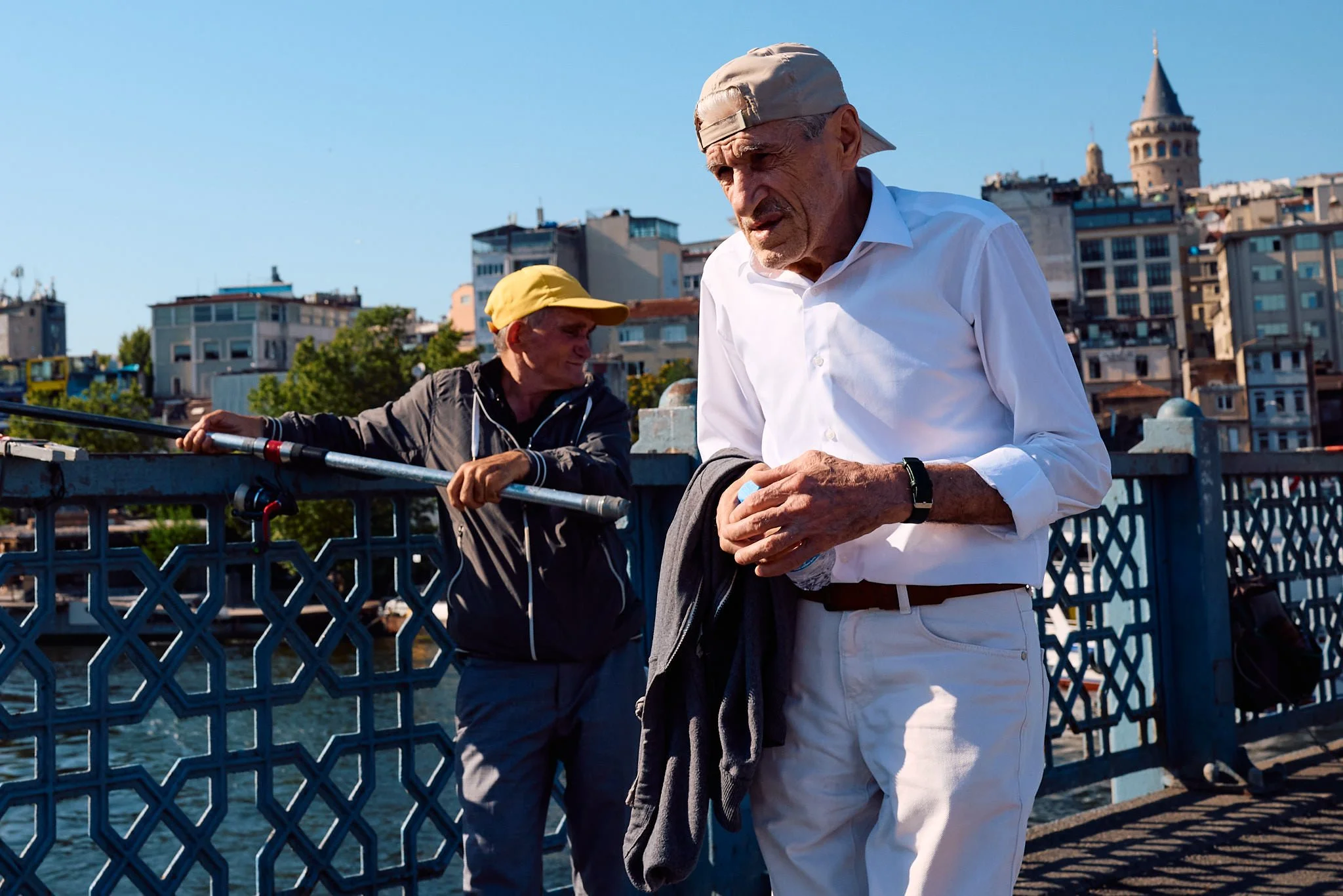 Older man in a white shirt and cap walking past a fisherman with his rod on the Galata Bridge, Galata Tower behind them