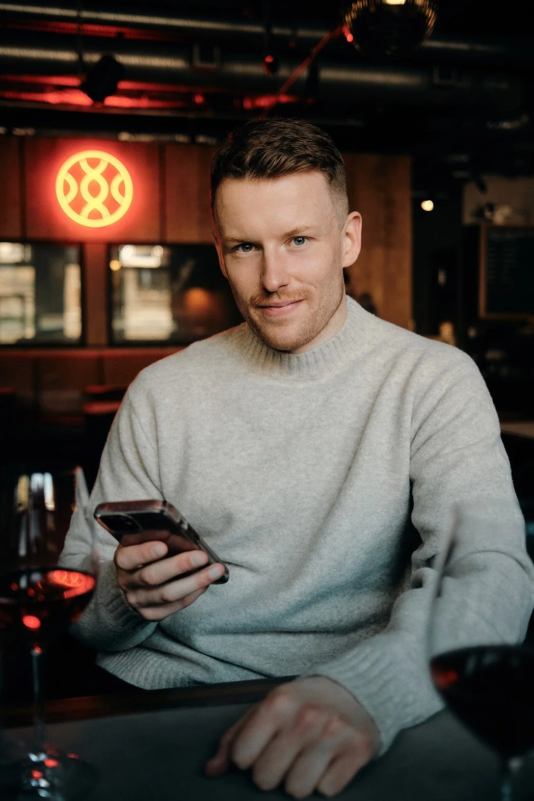 Dating photographer London lifestyle portrait of man looking at camera across table with phone in hand, slight smile, and neon light in background with reflection in glass of wine, relaxed and natural.