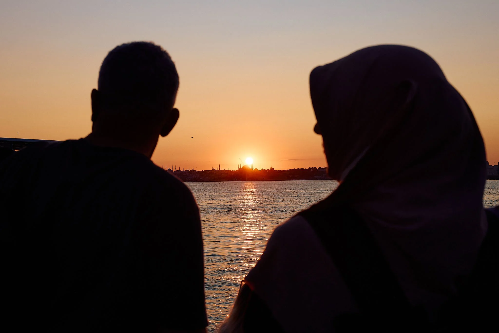 Silhouettes of a man and a woman in a headscarf watching the sun set over Istanbul’s old city skyline from the water