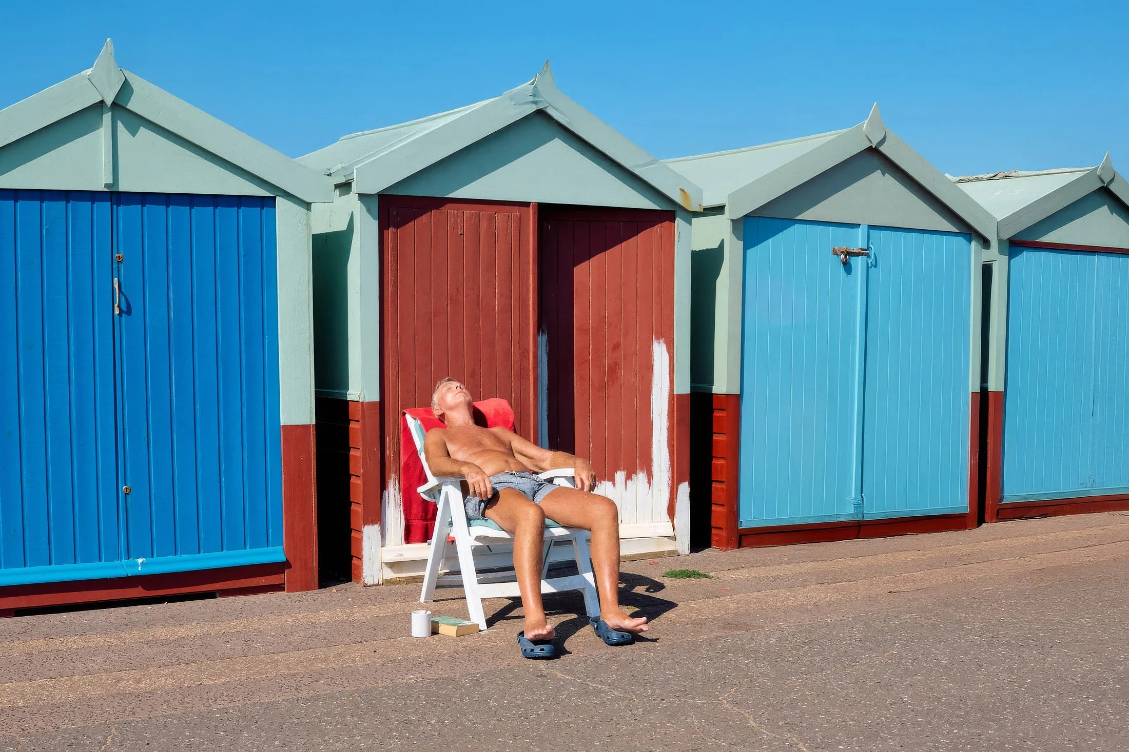Candid street photograph of a man sunbathing outside brightly painted red and blue beach huts beneath a clear sky.