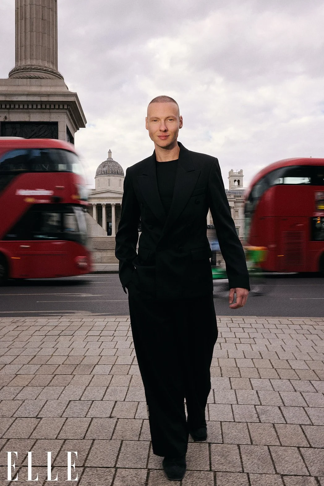 Personal branding photography of fashion consultant Bernard Garby walking toward camera with red bus, Lime bike, and Trafalgar square in background for ELLE Lithuania print magazine editorial
