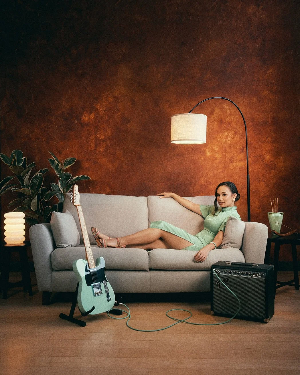 Personal branding photographer London portrait of music artist Denquar laying grey sofa wearing green dress and matching guitar with orange wall and plants.