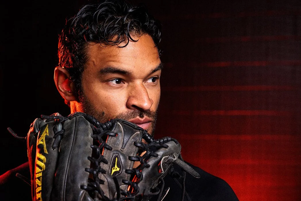 Advertising photographer London commercial lifestyle photography of a baseball coach posing in studio with glove over face, red light background on black brick wall with mesh for a public relations press release