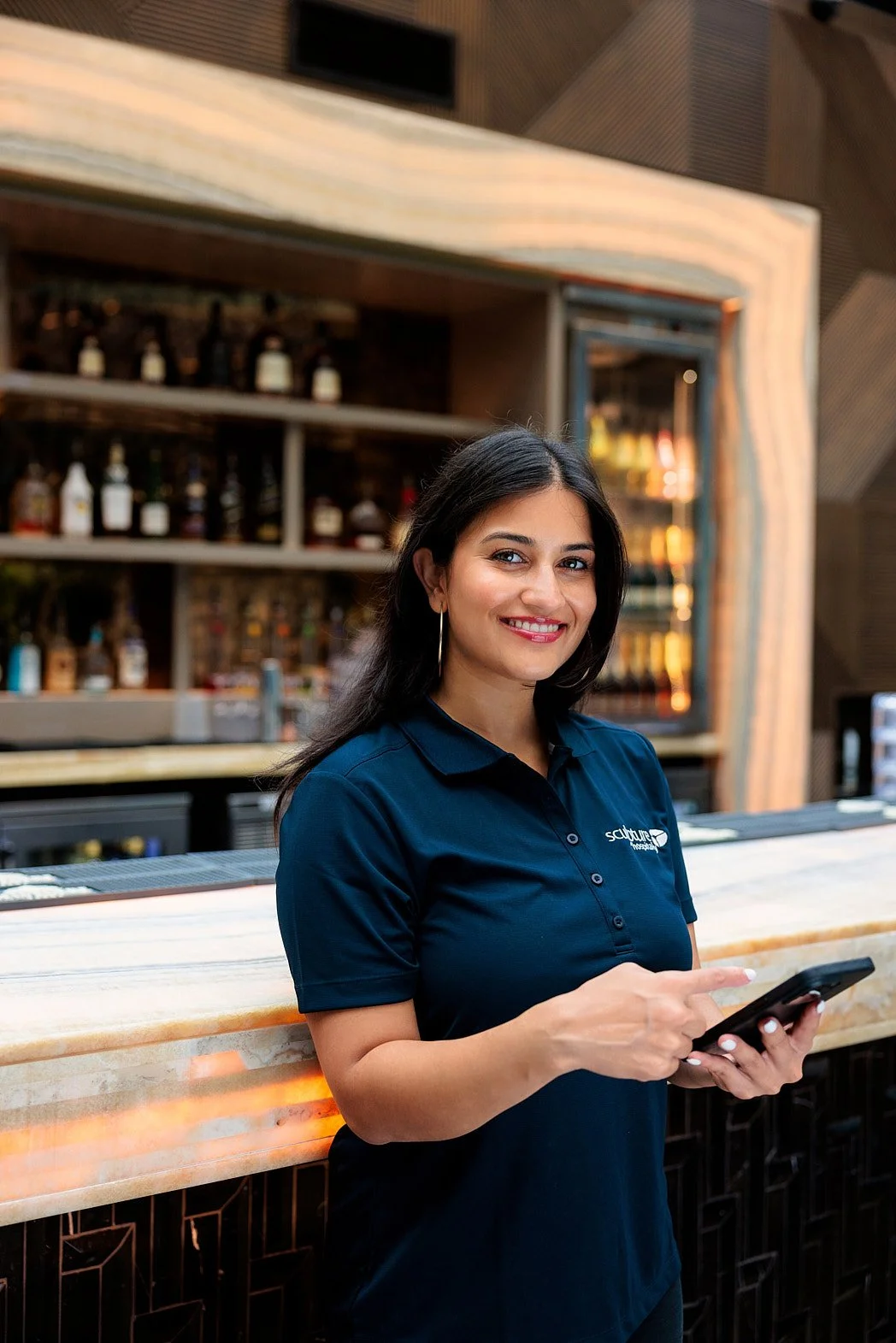 Advertising photographer London campaign image for bar inventory system of female manager in front of regal night club smiling at camera with bottles in background holding tablet.