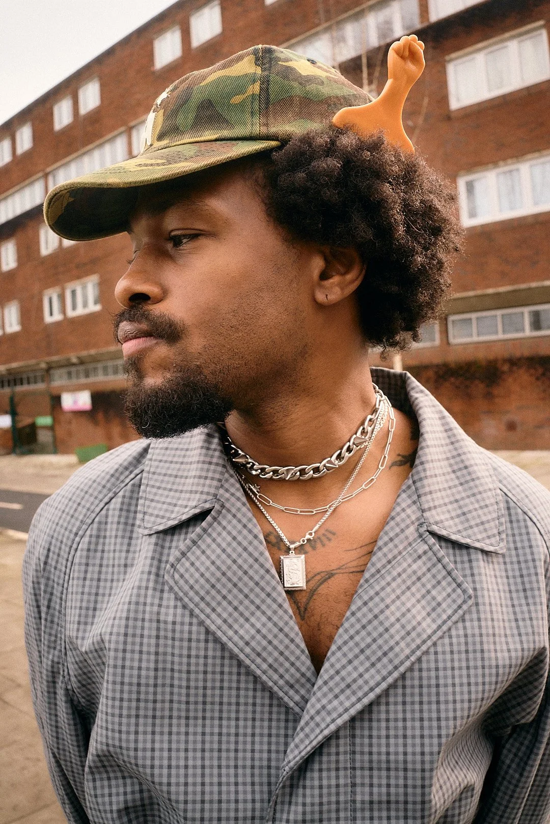 Street fashion portrait of music artist Duckwrth at London Fashion Week with plaid trench coat camo hat orange hair pick silver necklaces looking off