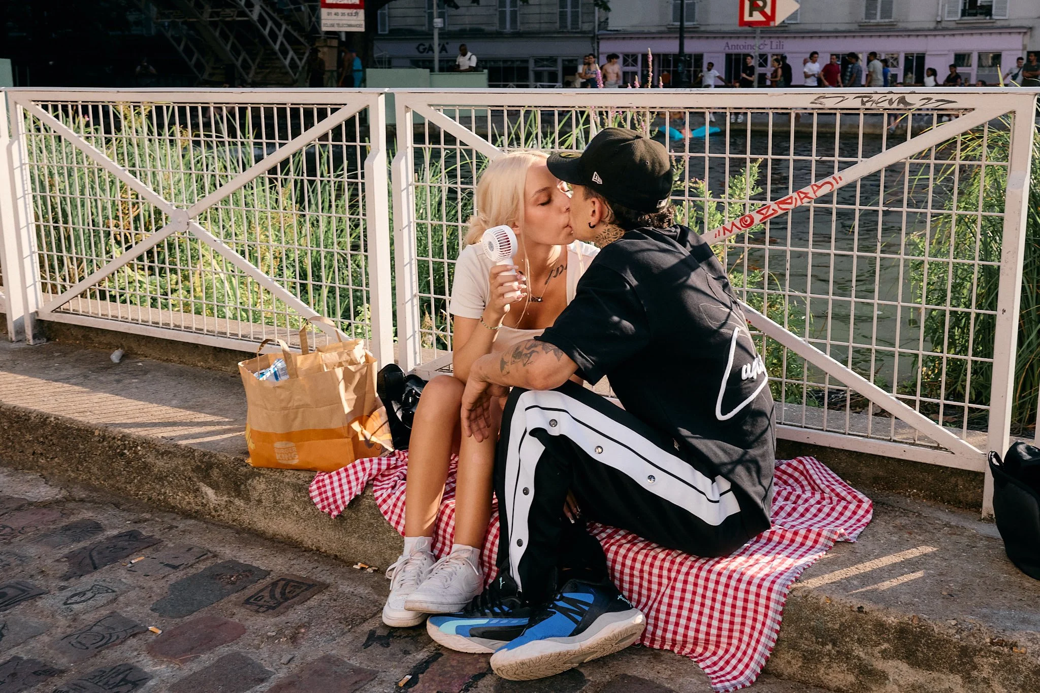 Couple kissing on a red gingham blanket by Canal Saint-Martin, the woman holding a white electric handheld fan in 40°C heat, Paris Fashion Week SS27 menswear