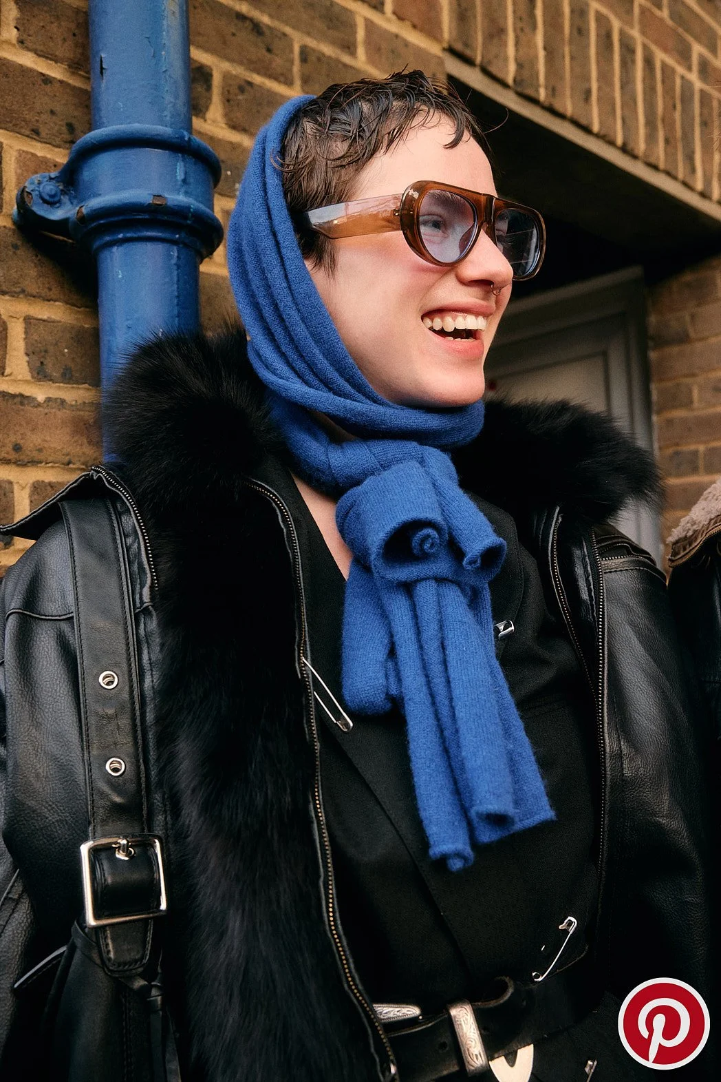 Advertising photography of campaign street style portrait at London Fashion Week documenting street fashion for Pinterest of woman wearing blue scarf over head with blue sunglasses looking off smiling blue and brick background