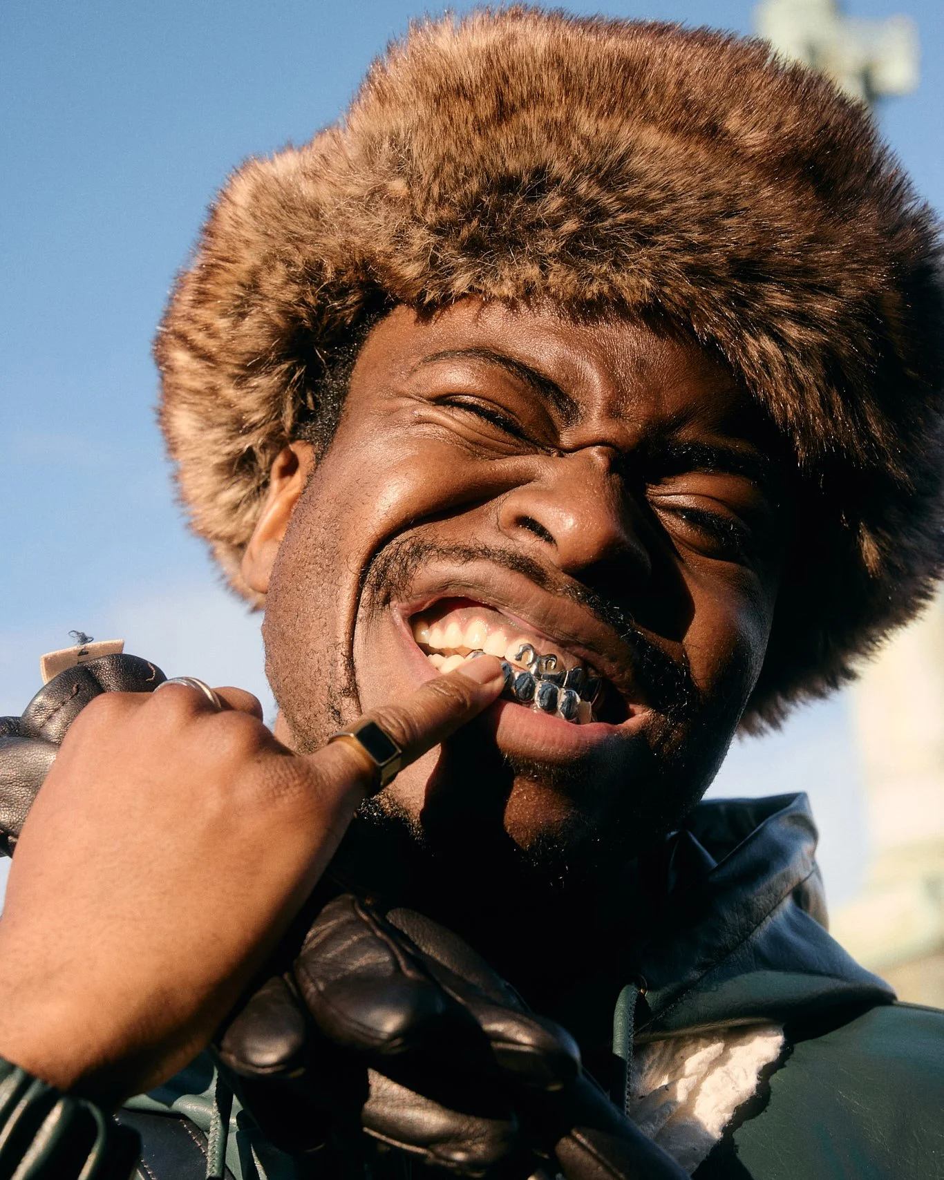 Street photography of street style at London Fashion Week of man showing teeth jewelery close up in sunlight wearing fur hat