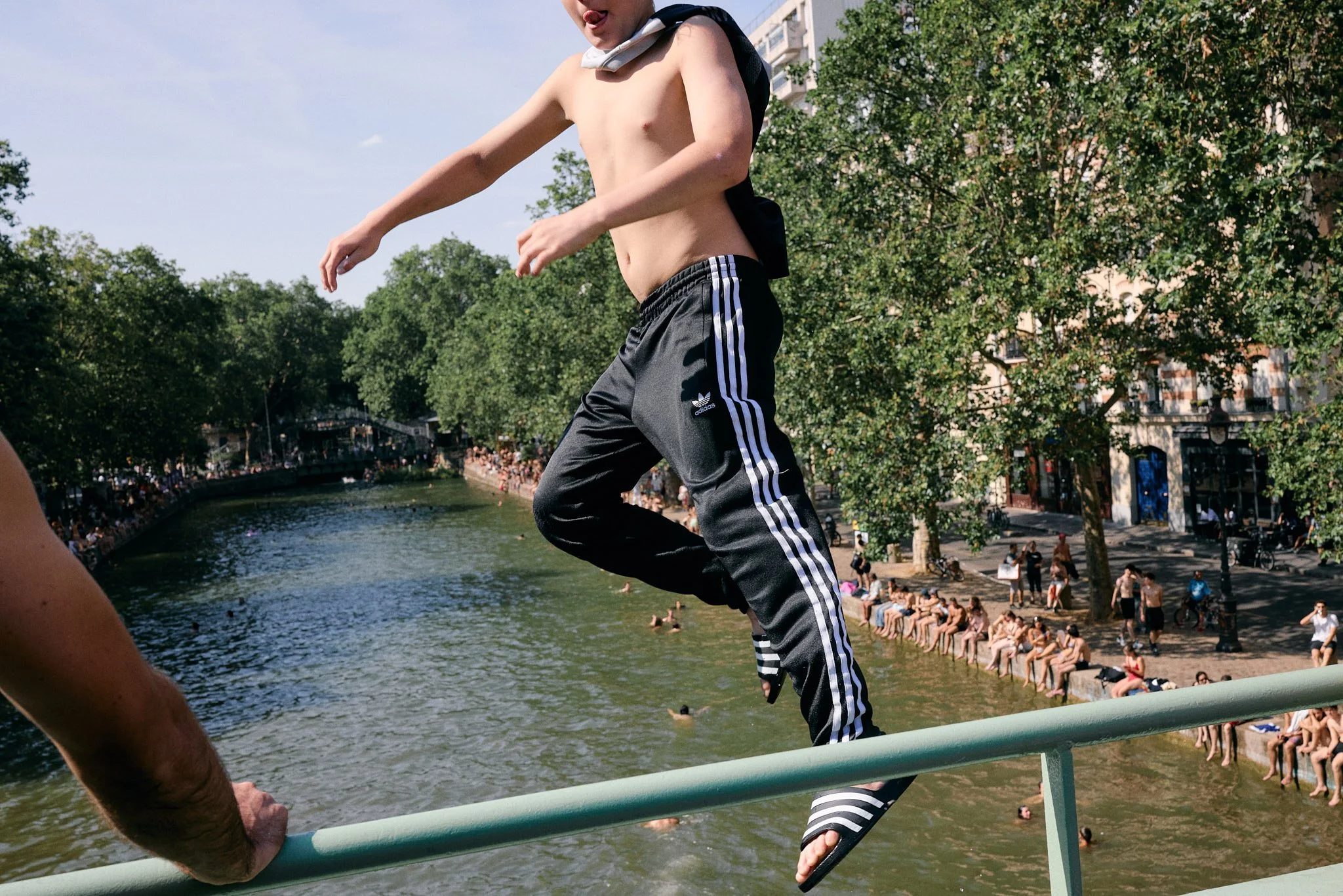 Boy in adidas trackpants and adidas slides frozen mid-jump above Canal Saint-Martin as crowds line the banks, Paris Fashion Week SS27 menswear