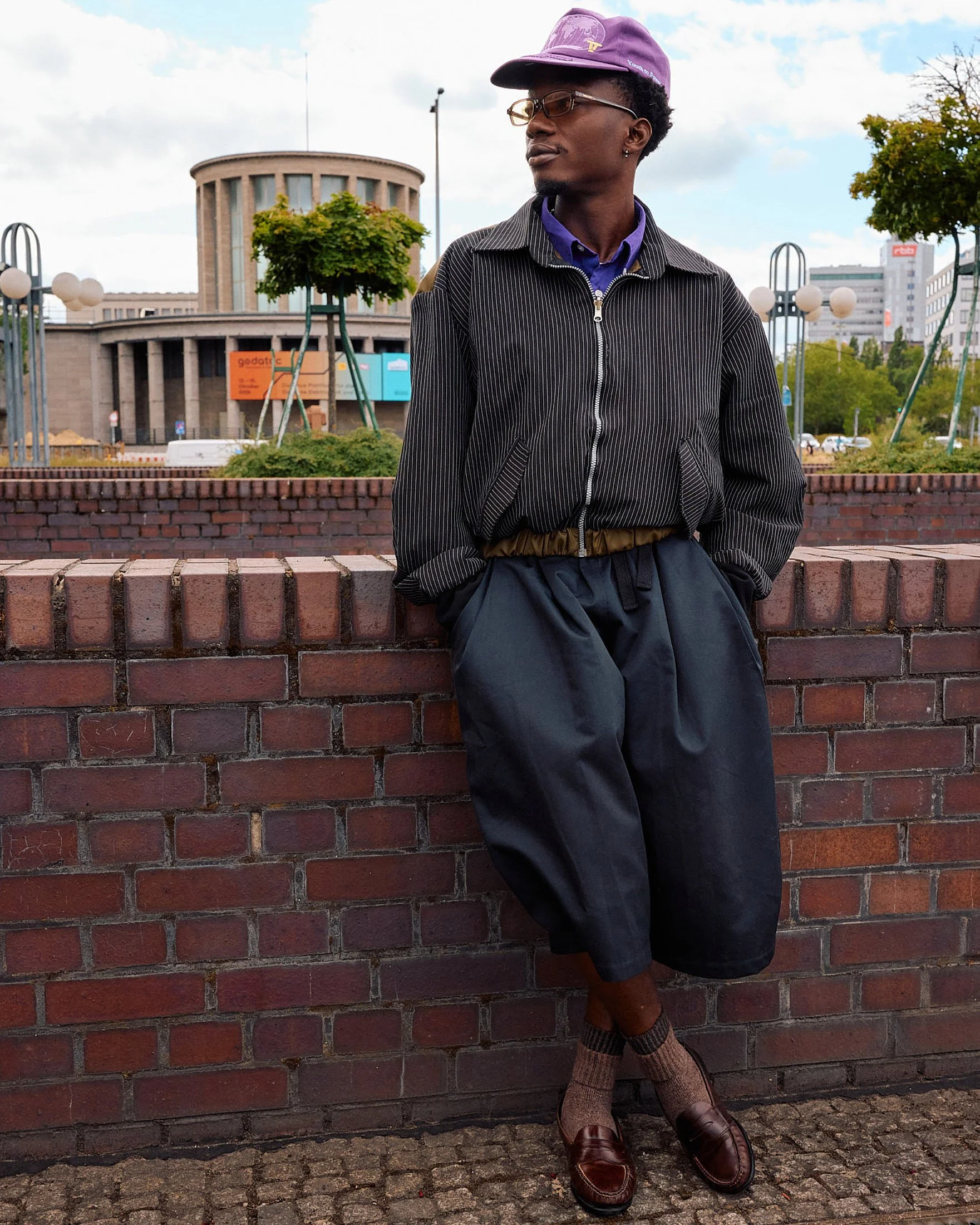 Menswear guest leaning on brick in a pinstripe zip blouson over a purple shirt, wide pleated navy culottes, ribbed socks and burgundy penny loafers, Berlin Fashion Week SS27