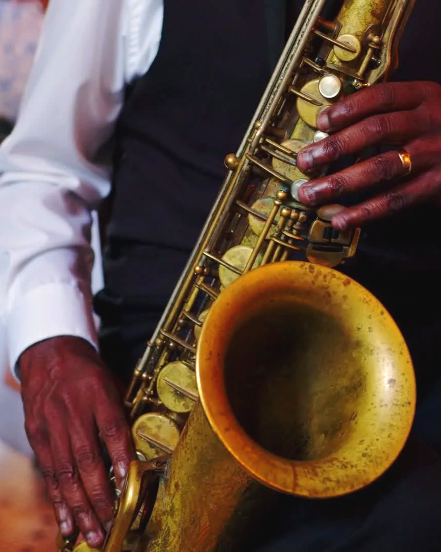 Close-up of a person playing a vintage gold-colored saxophone, with their hands on the instrument and wearing a wedding band.