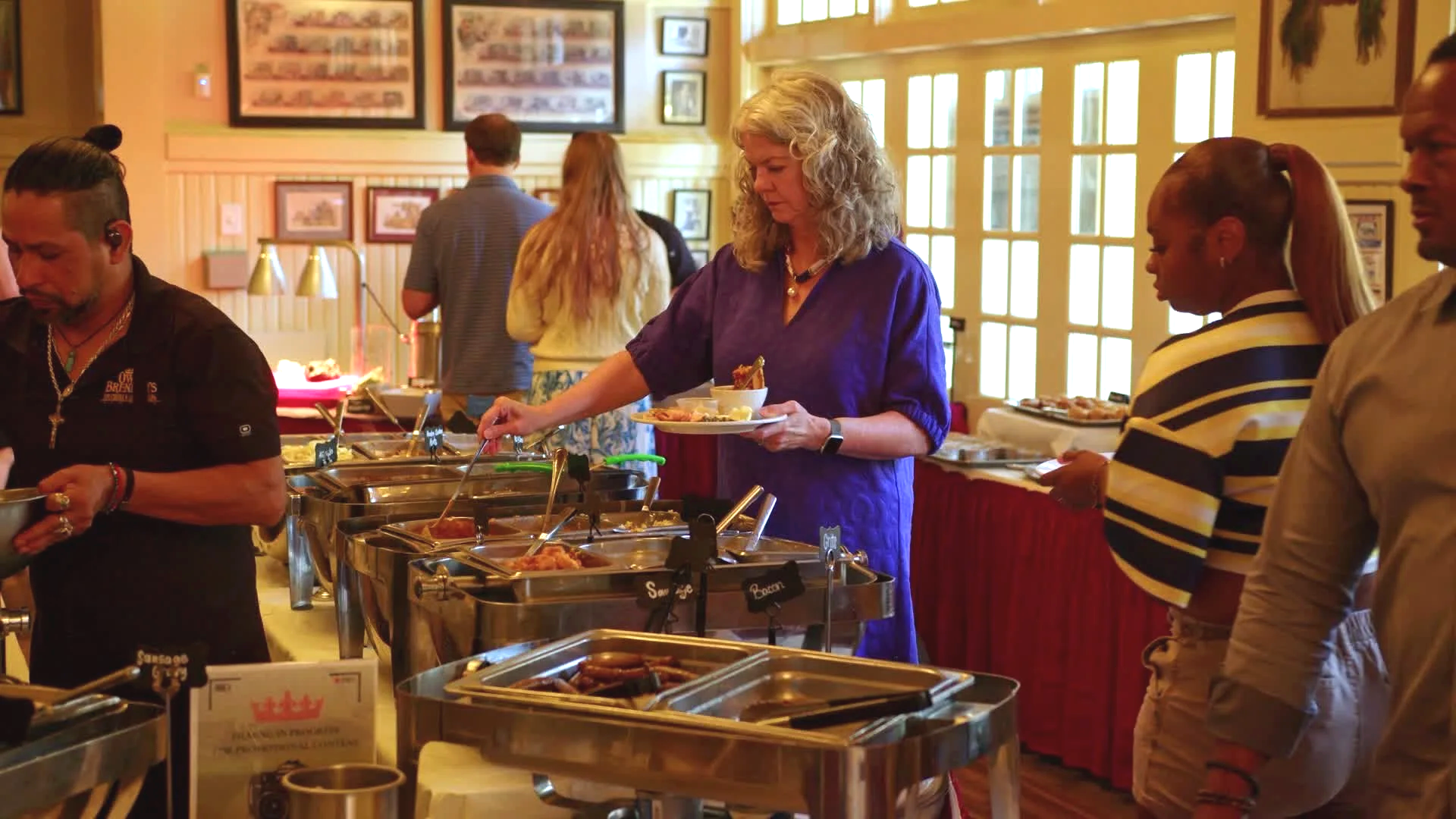 People serving themselves food at a buffet in a dining area with wooden walls and large windows.