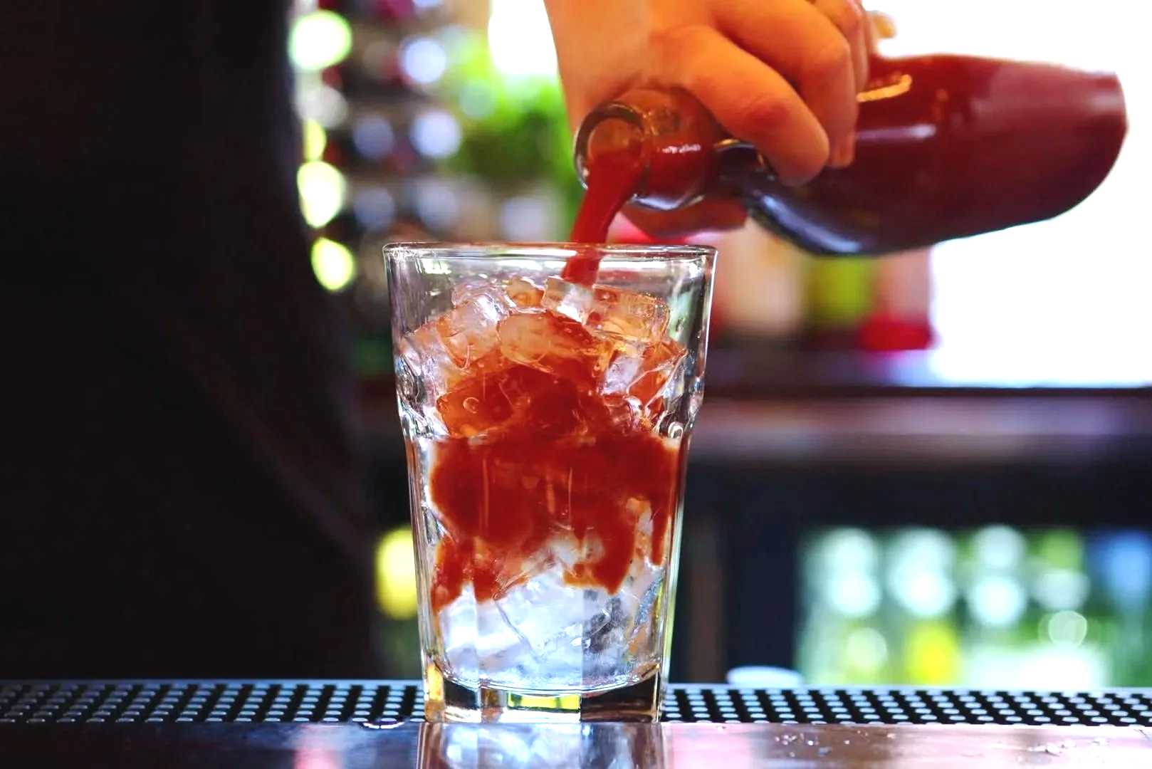 A hand pouring a red liquid from a glass bottle into a glass of ice cubes.