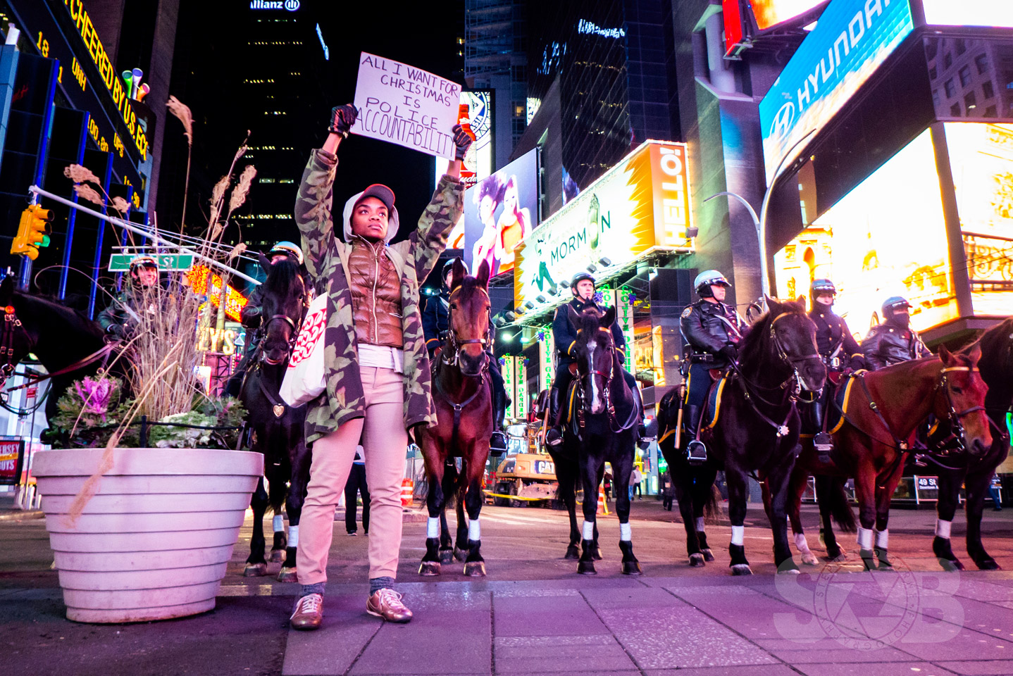 Protester in Times Square holding a sign in front of several riot police on horse back.