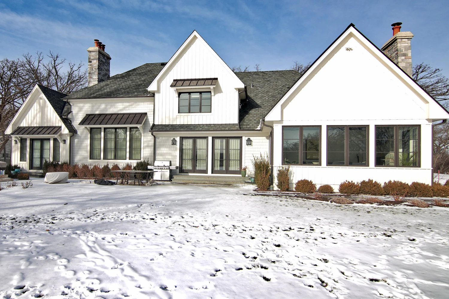 White house with multiple gables, large windows, and chimneys, surrounded by snow on the ground, with leafless trees in the background.