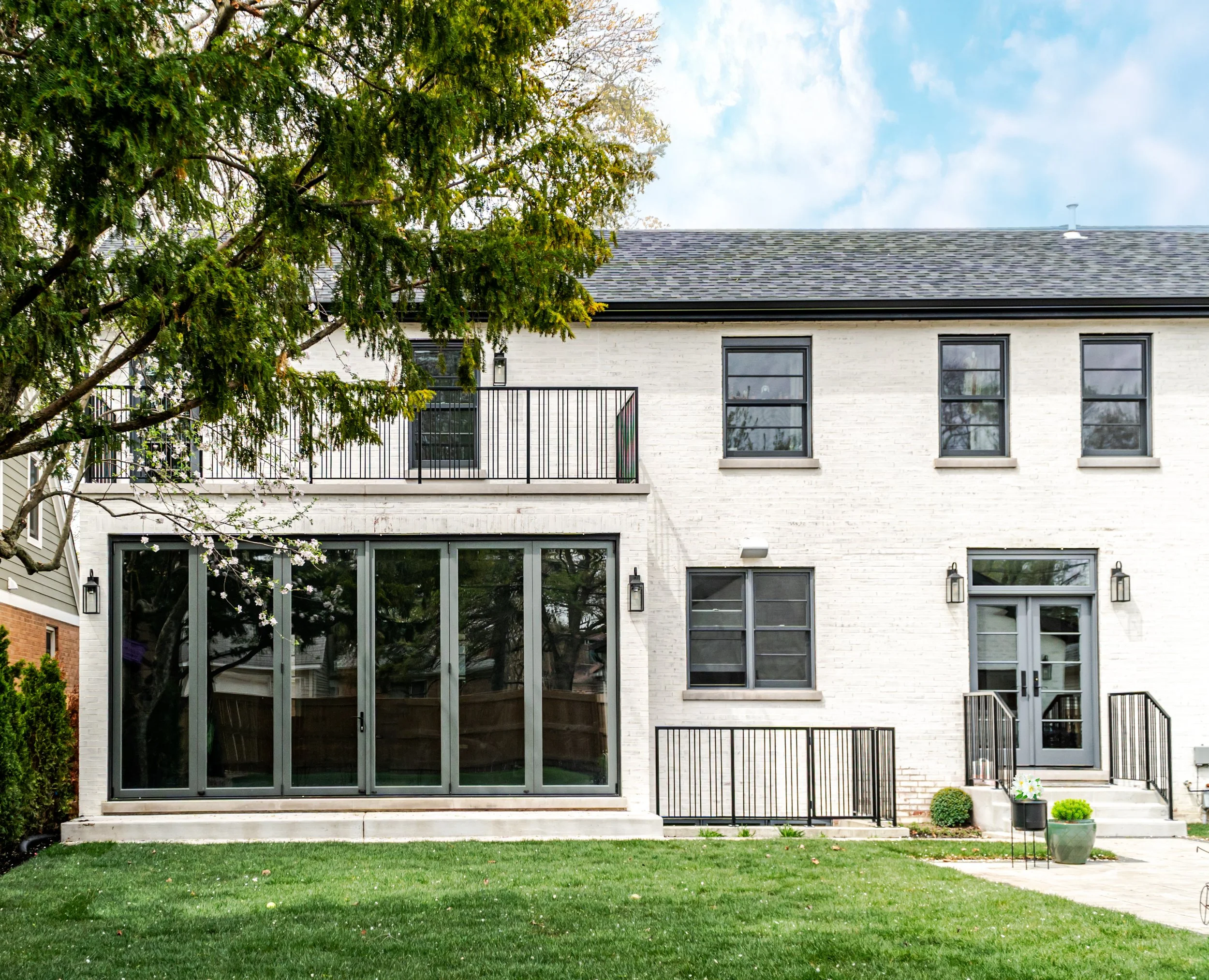 Back of a modern white brick house with large glass sliding door, several windows, a small balcony with black railing, and a well-kept lawn with potted plants.