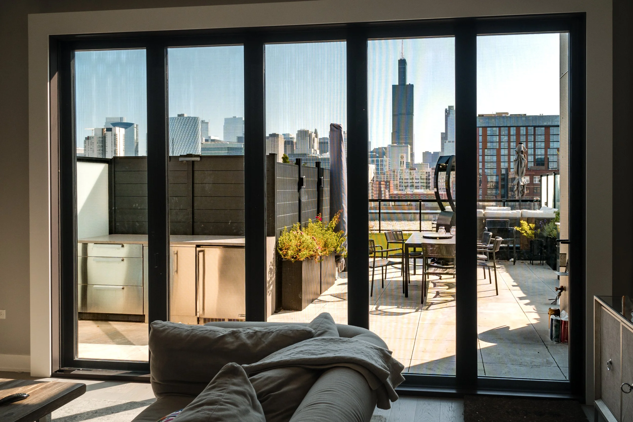 View of a city skyline with tall skyscrapers from an apartment balcony, seen through sliding glass doors, with outdoor furniture and plants on the balcony.