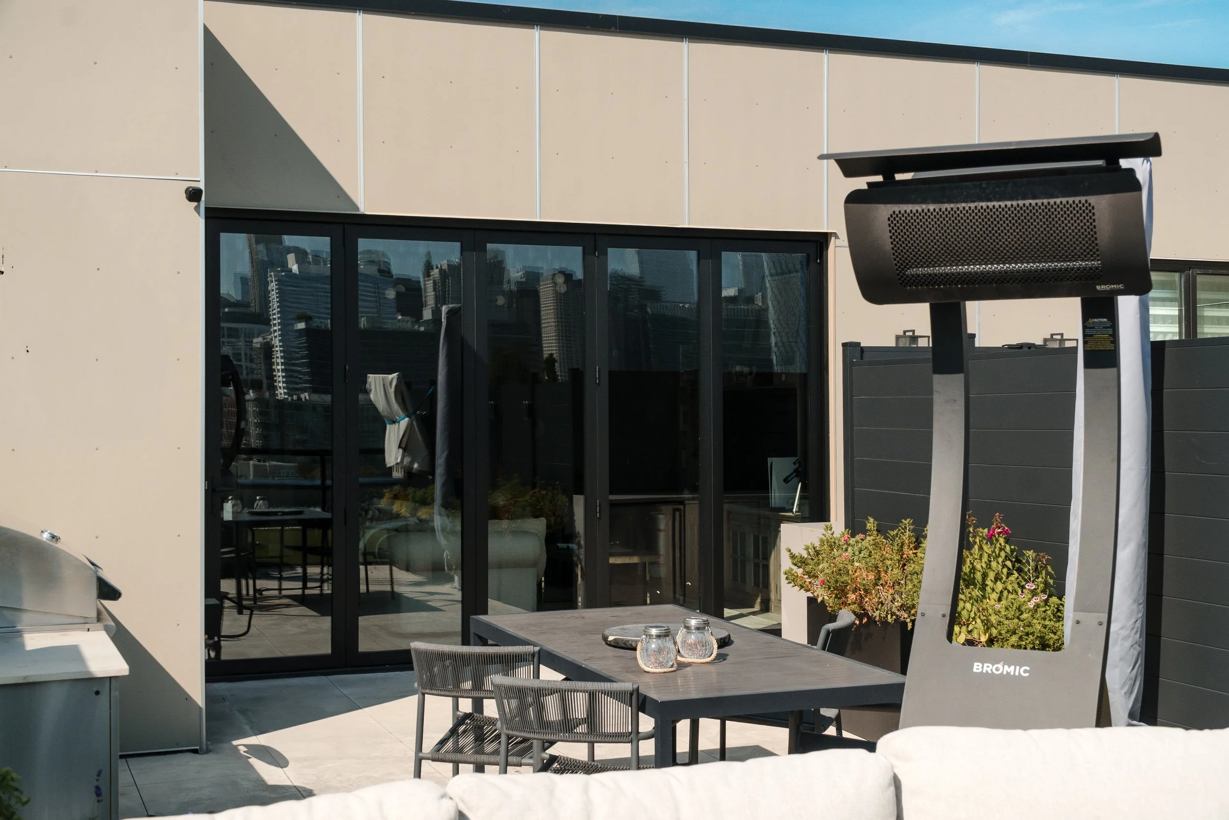 An outdoor patio with a table and chairs, a large black heater, and a glass sliding door leading inside a modern apartment or building.