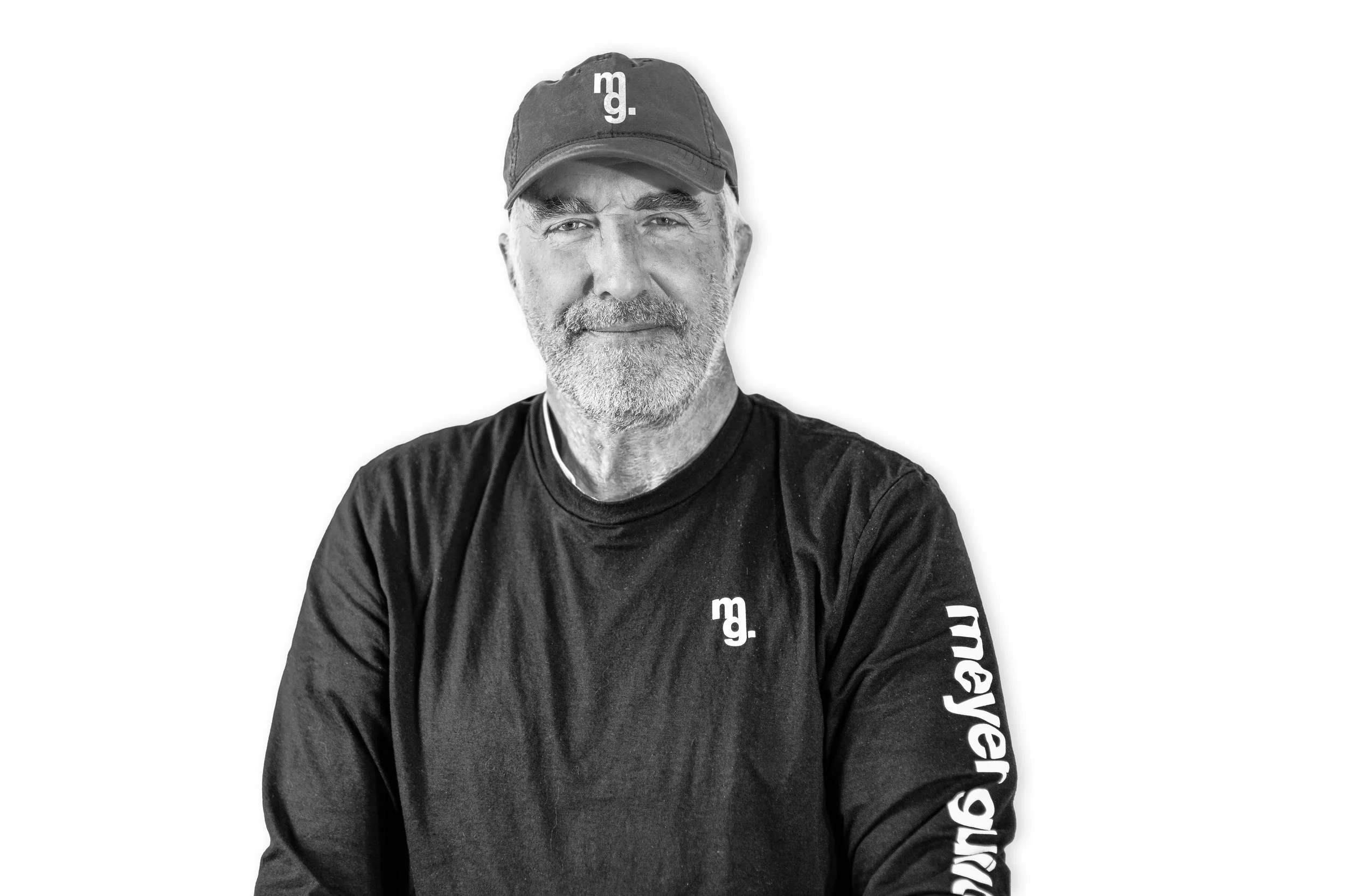 Black and white portrait of an older man with a beard, wearing a baseball cap and a long-sleeve shirt with logos, against a plain background.