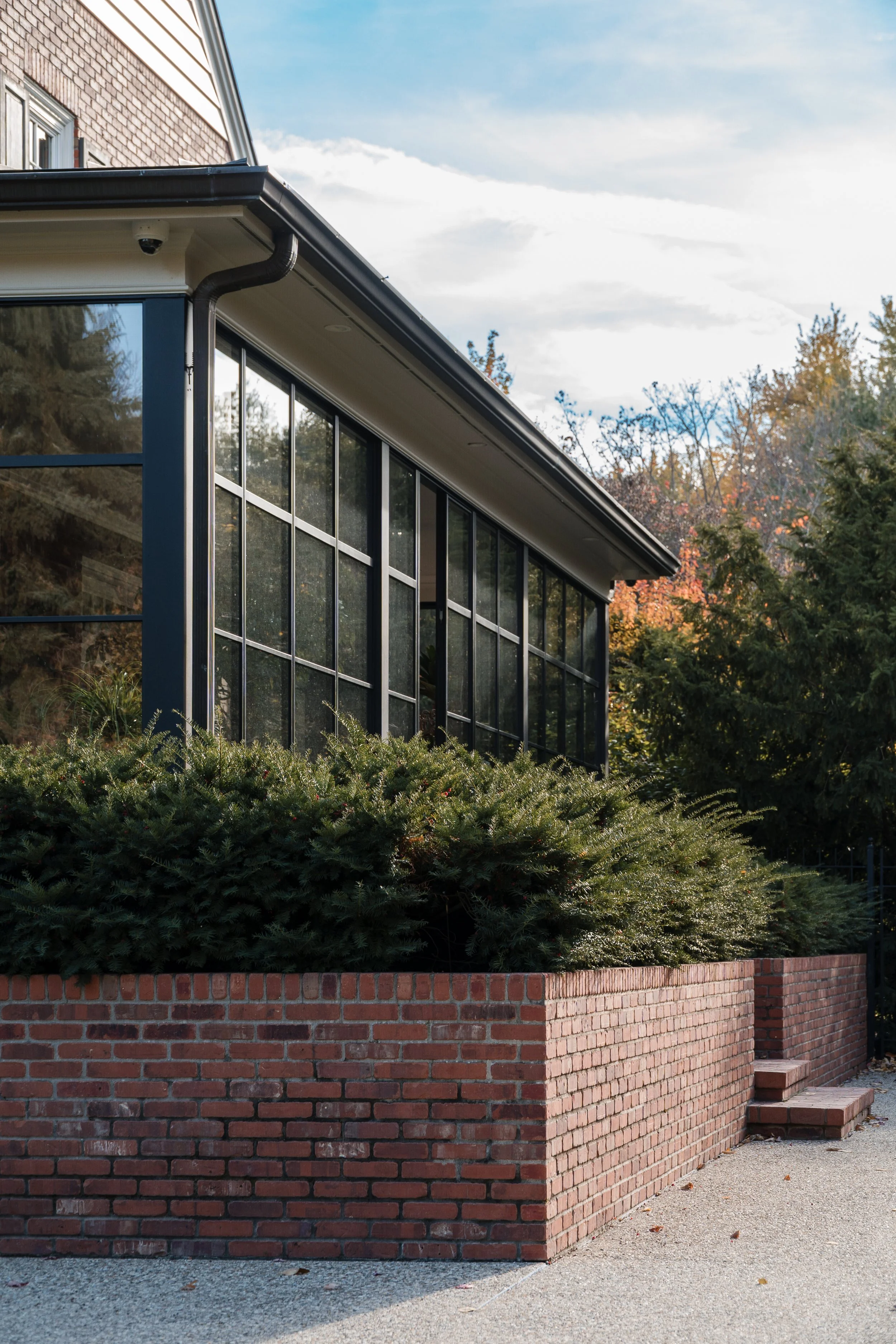 Modern building with large glass windows, brick planter with bushes, and steps leading up to a door, surrounded by trees in fall.