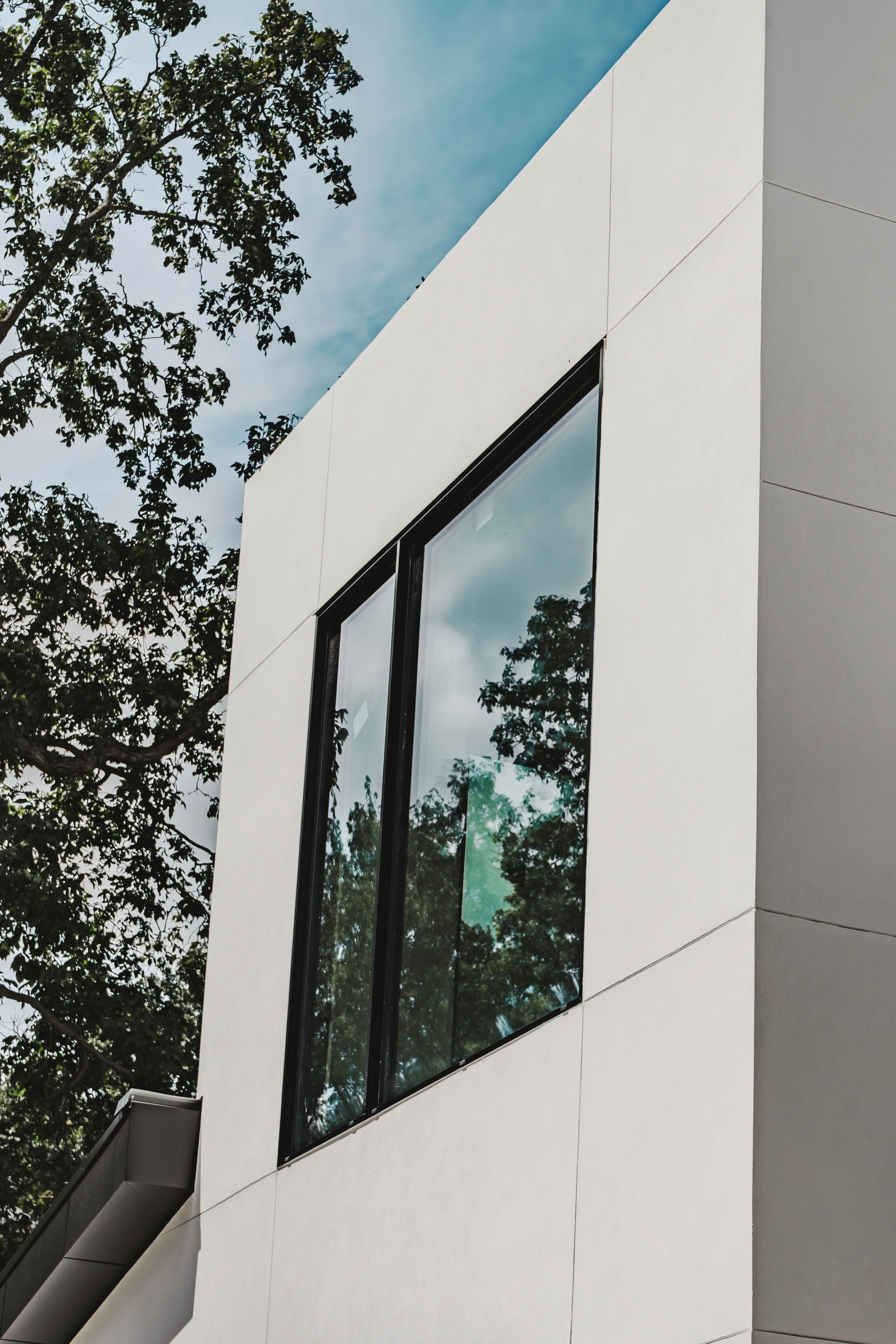 Close-up view of a modern white building with large reflective windows, surrounded by trees with a partly cloudy sky above.