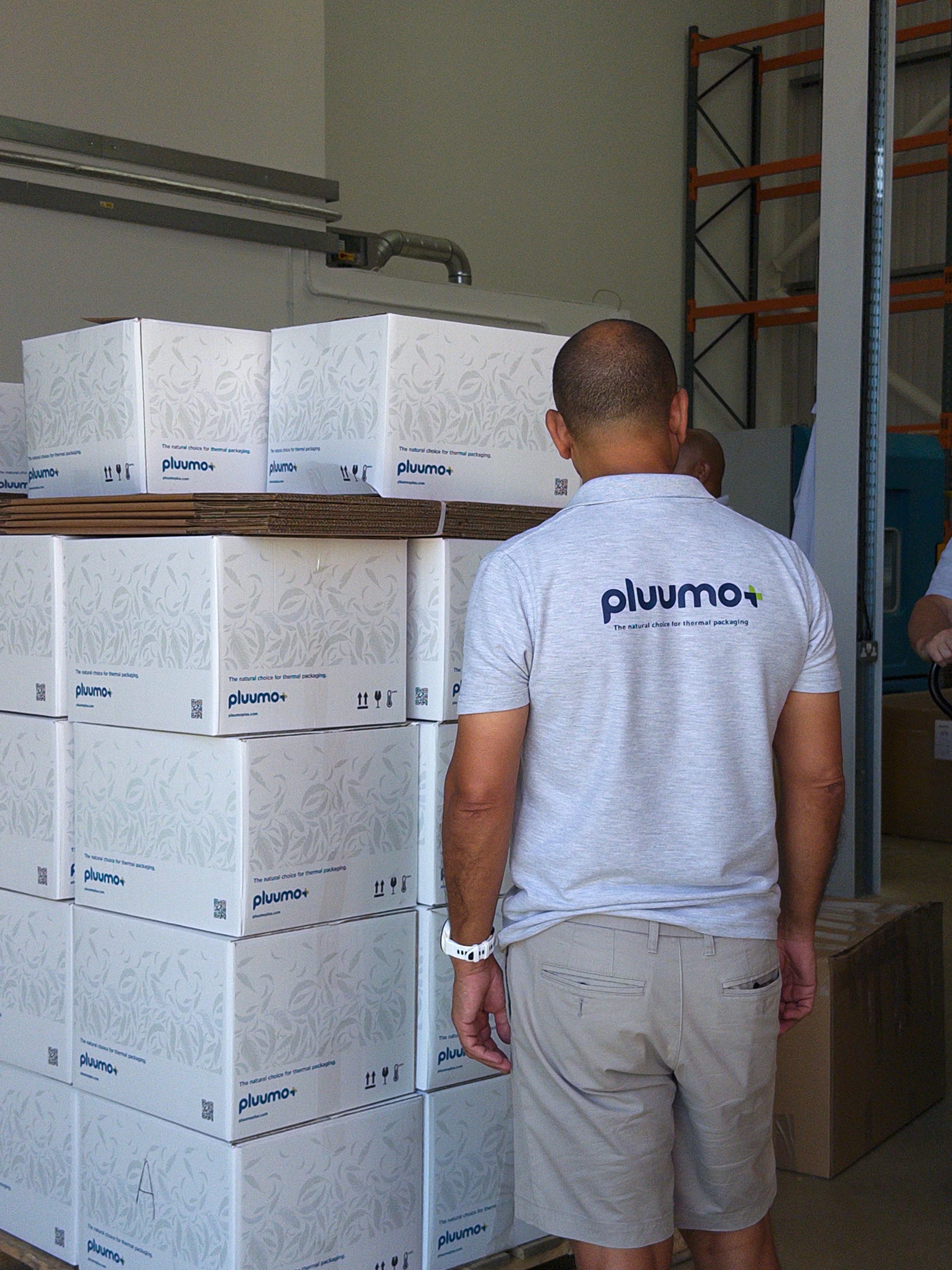 A man and a woman in a warehouse or storage area, viewing boxes labeled 'pluumo+' with the tagline 'The natural choice for thermal packaging.' The man has a shaved head and is wearing a gray polo shirt and beige shorts. The woman is partially visible