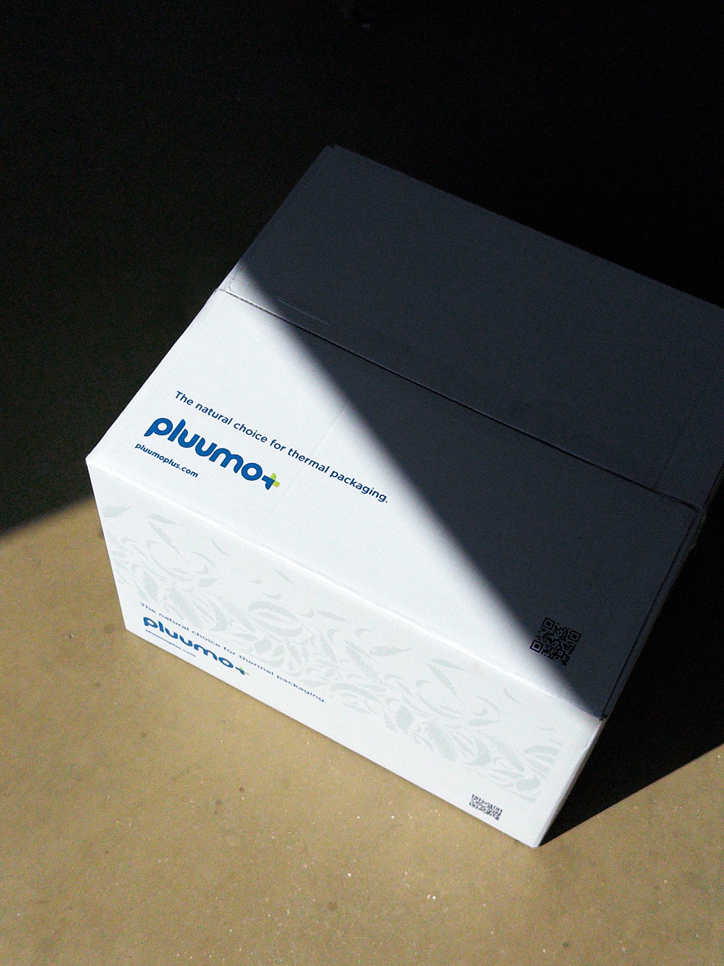 A white box with blue and green text on a beige surface, partially in shadow. The box has branding for "Plum" and mentions thermal packaging.