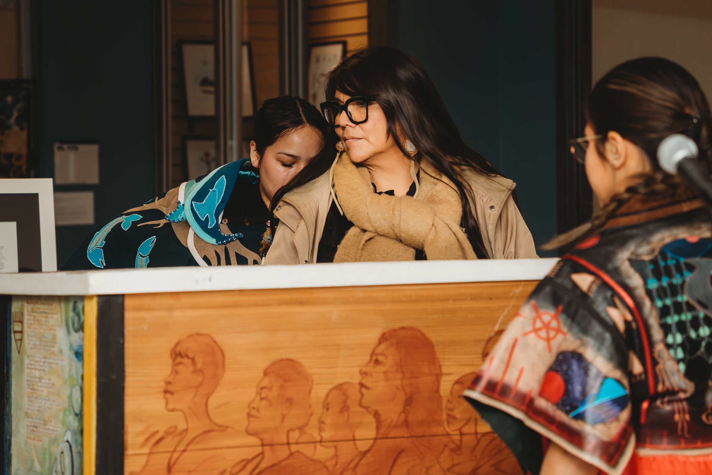 An Indigenous Artist (female) stands behind a bentwood box designed by multiple artists, representing the four colours of the Medicine Wheel. She is surrounded by two Indigenous women in regalia.