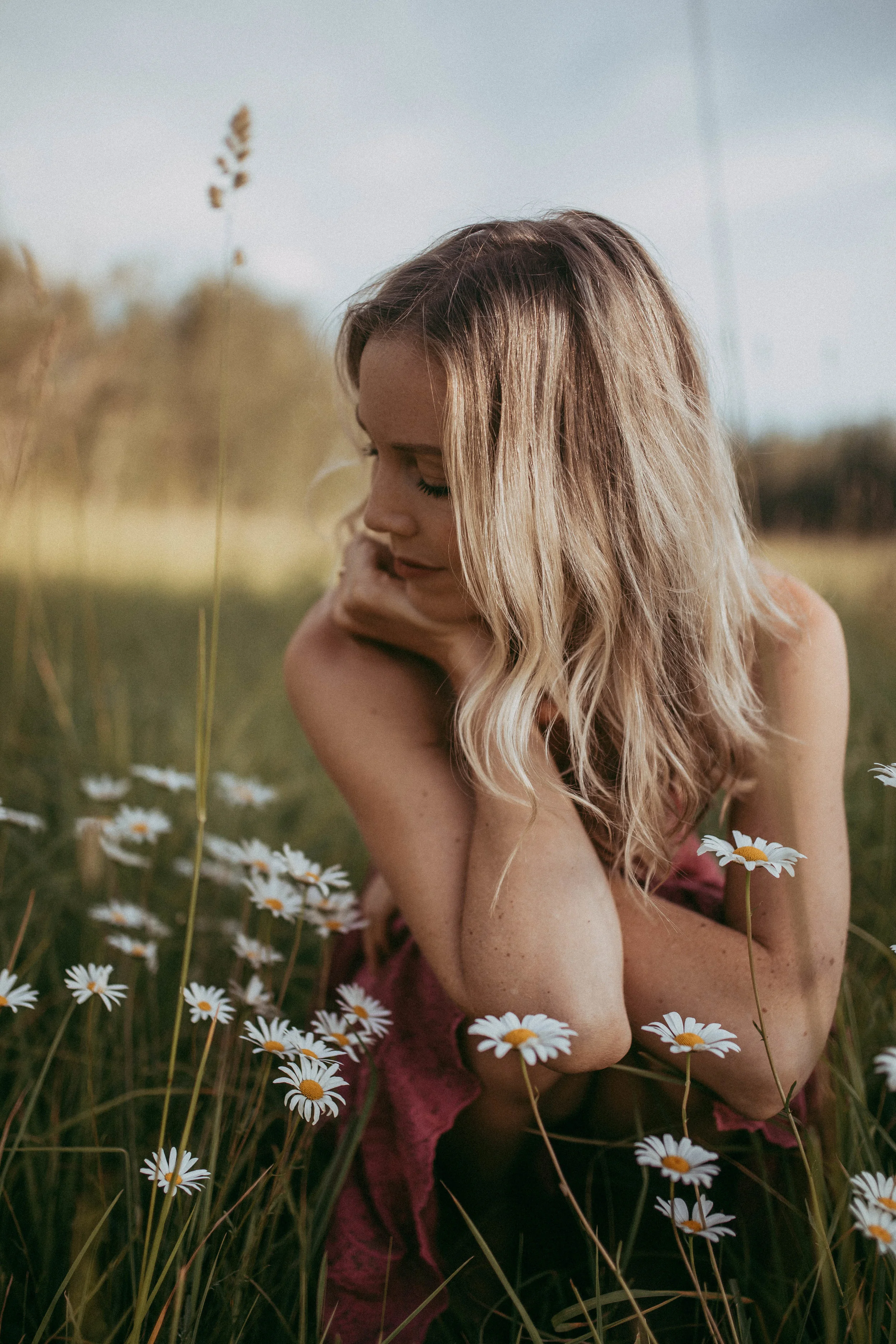 Woman with blonde hair sitting in a field of daisies, looking down, surrounded by greenery and wildflowers.