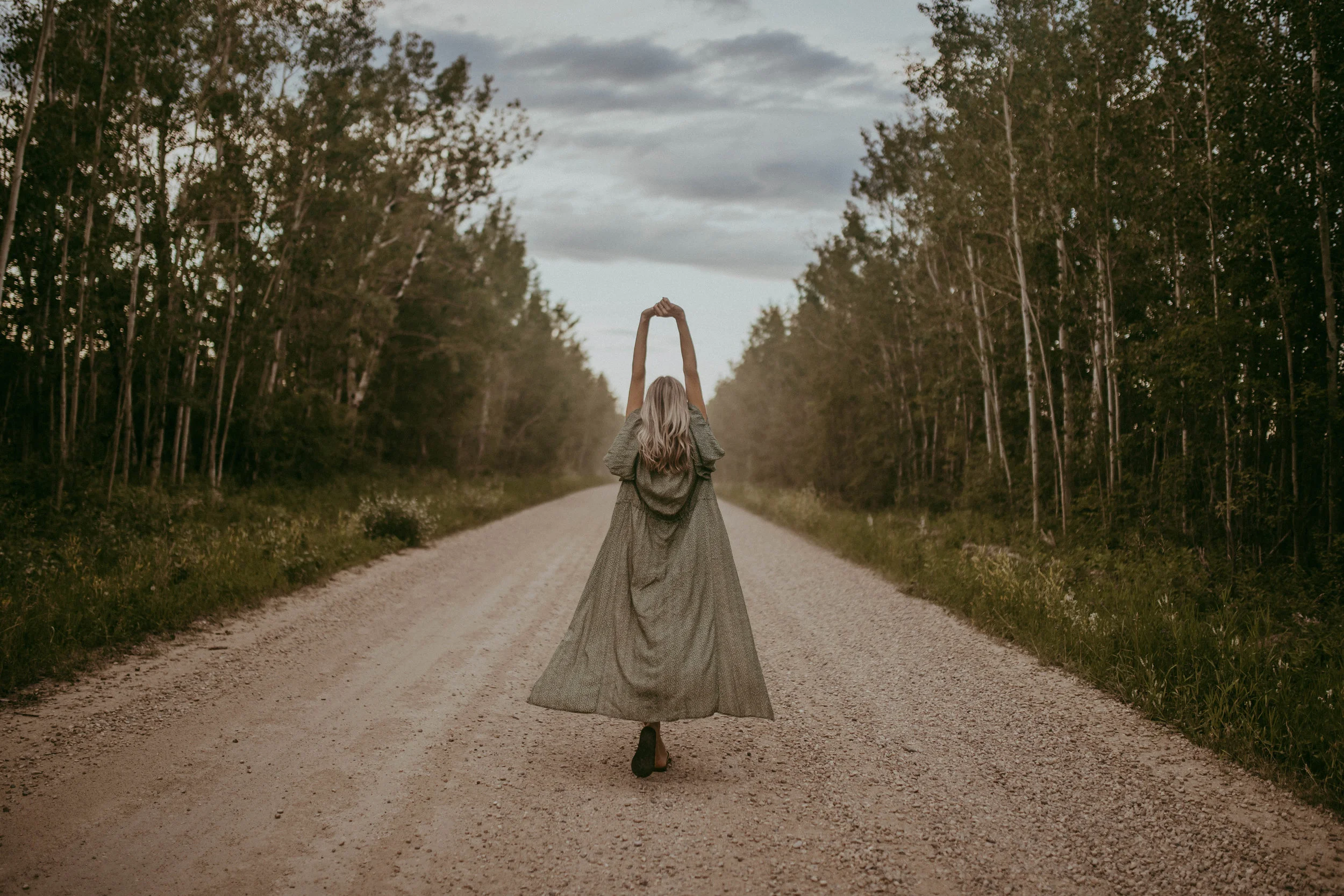Person in a long dress standing on a gravel road with arms raised, surrounded by trees under a cloudy sky.