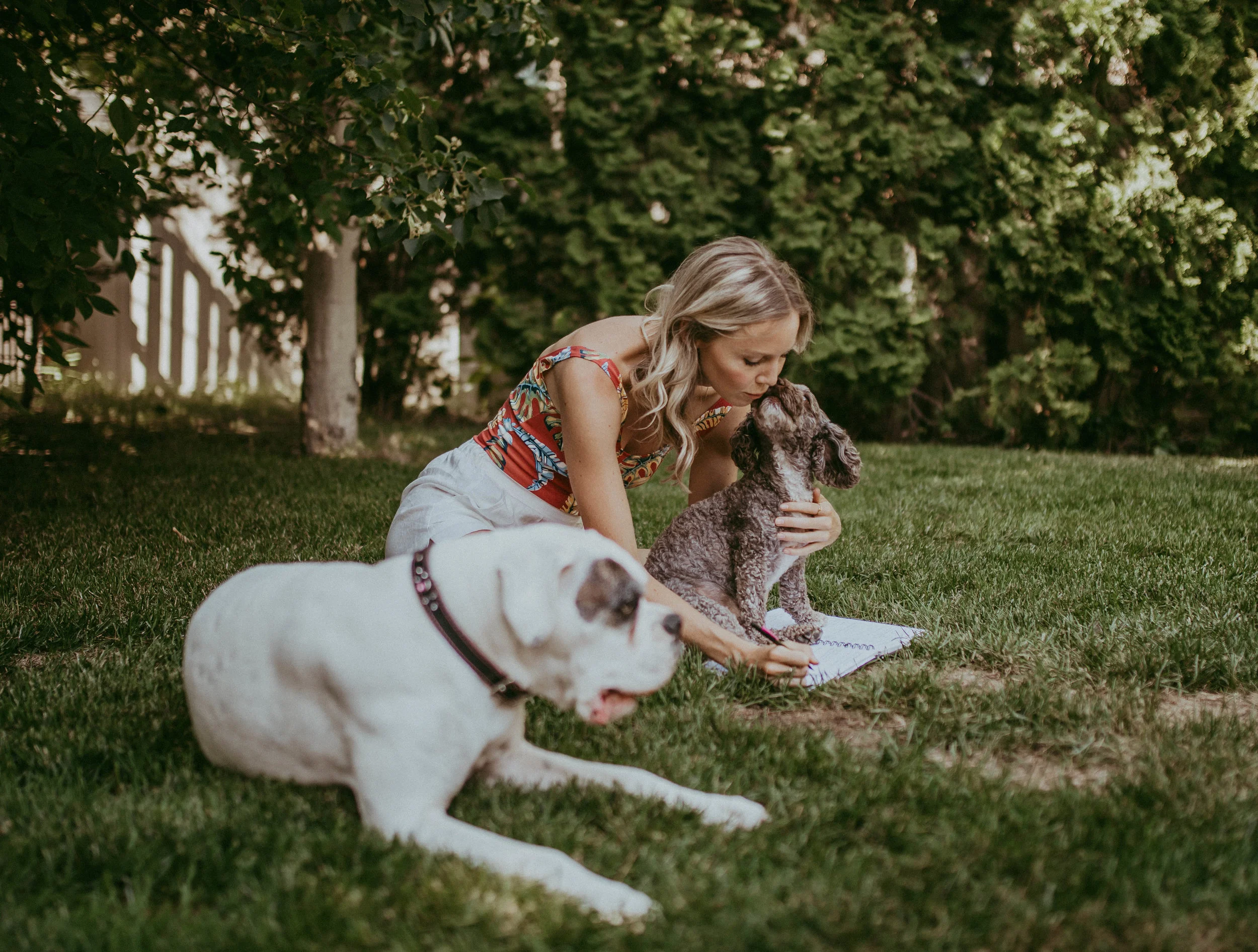 A woman kneeling on grass, kissing a small dog while holding a book, with another larger dog lying nearby. Trees and a fence are in the background.