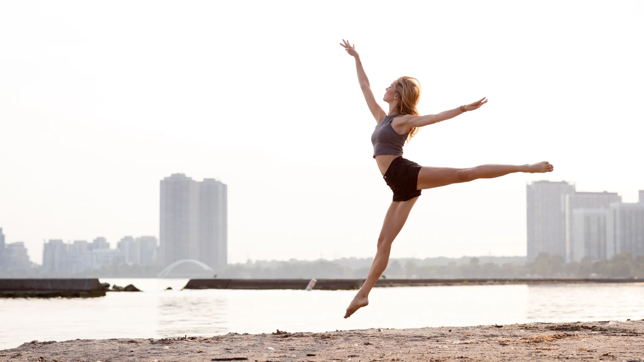 Woman doing a ballet leap at the beach with city skyline in background.