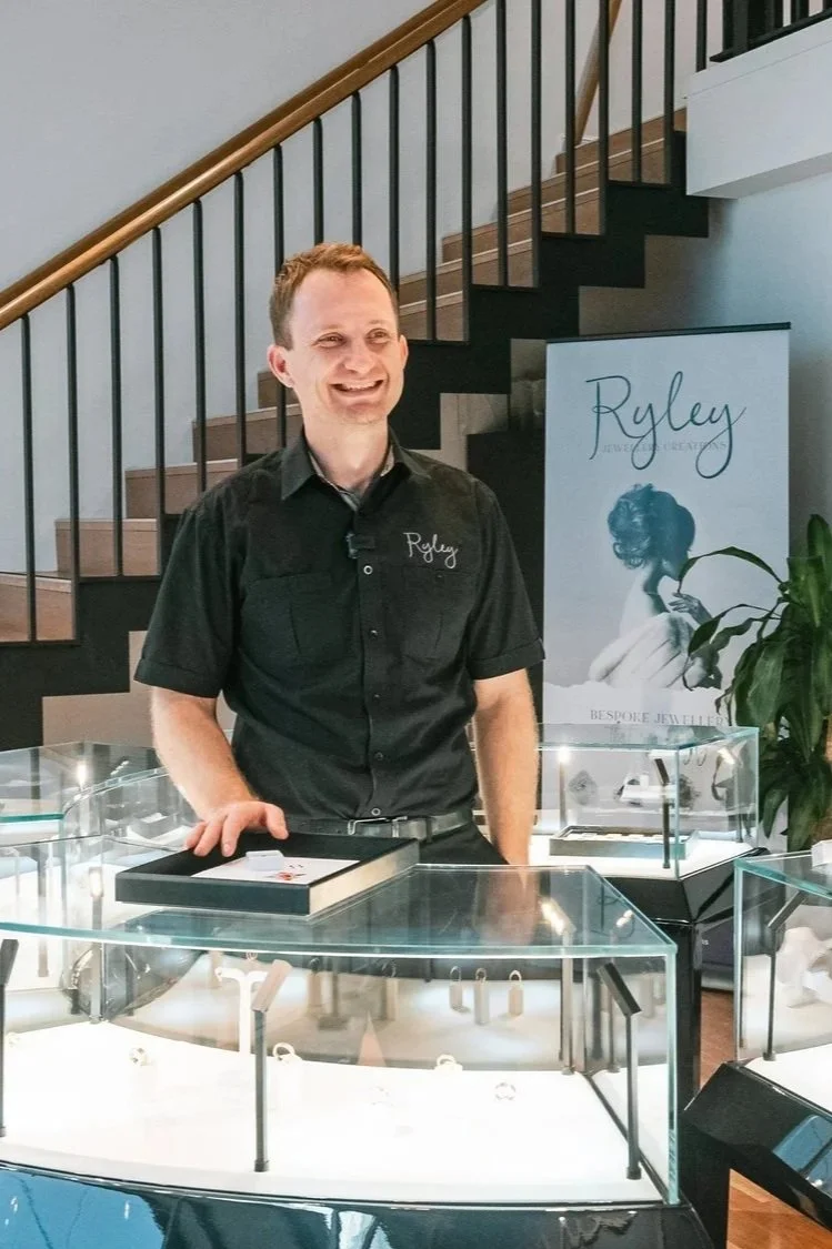Ewen Ryley wearing a black shirt with the name 'Ryley' embroidered on it, standing behind a glass display case filled with jewelry, smiling at an indoor event with a staircase and a promotional poster in the background.