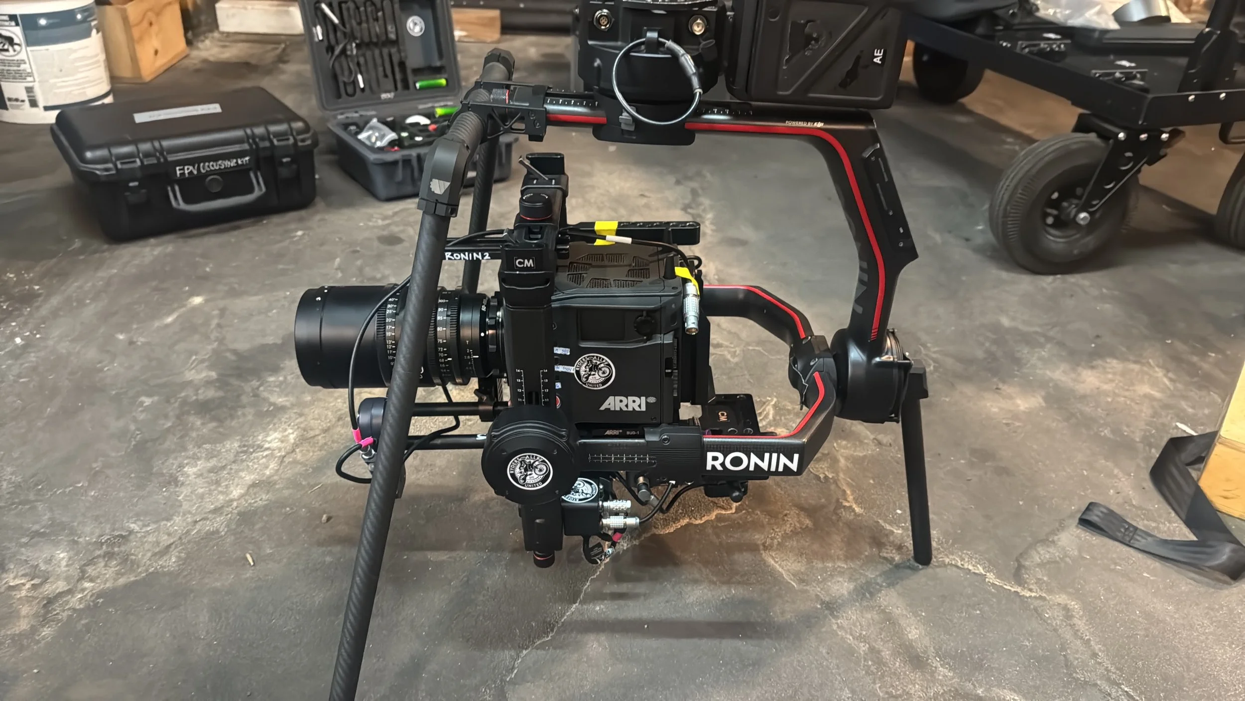 A professional camera mounted on a Ronin camera stabilizer rig with various equipment cases and tools in the background, on a concrete floor in a workspace.