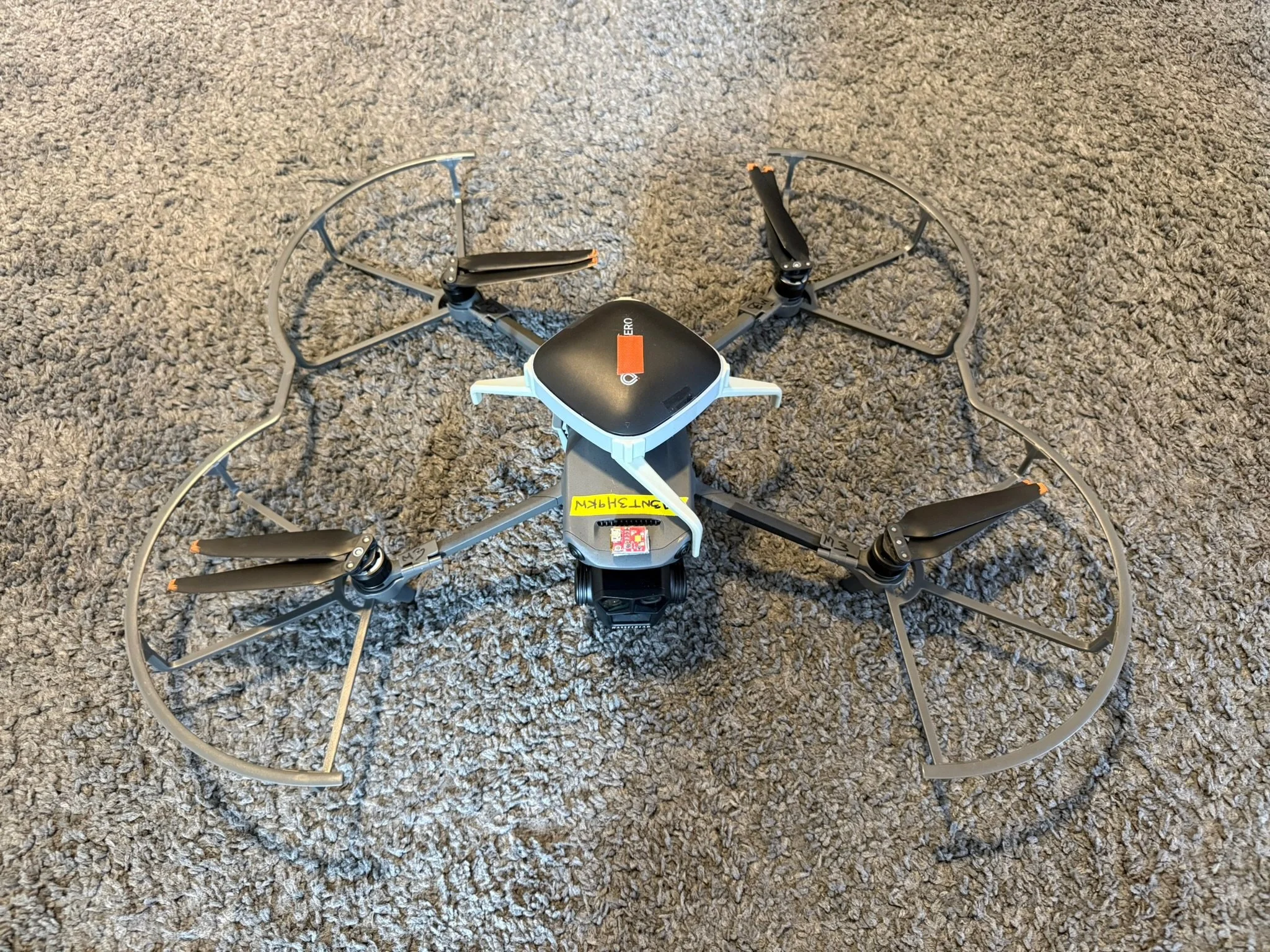 A quadcopter drone with four propellers and a protective cage, resting on a gray carpeted surface.