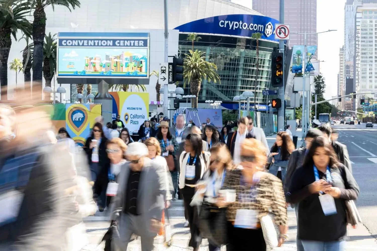 A large crowd of attendees walking across the pedestrian plaza from Crypto.com Arena toward the Los Angeles Convention Center for a major summit.