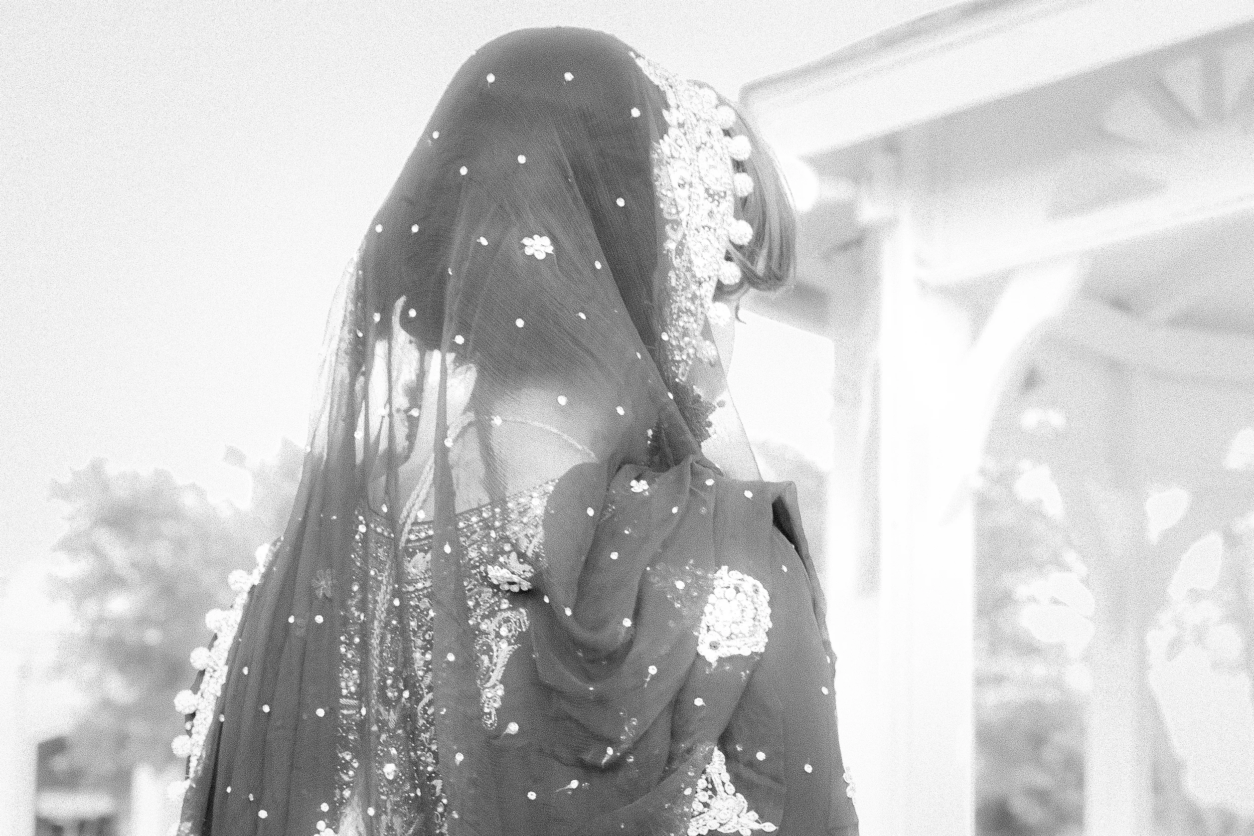Black and white image of a person wearing an elaborate, beaded veil and traditional attire, possibly a sari, standing outdoors next to a structure.