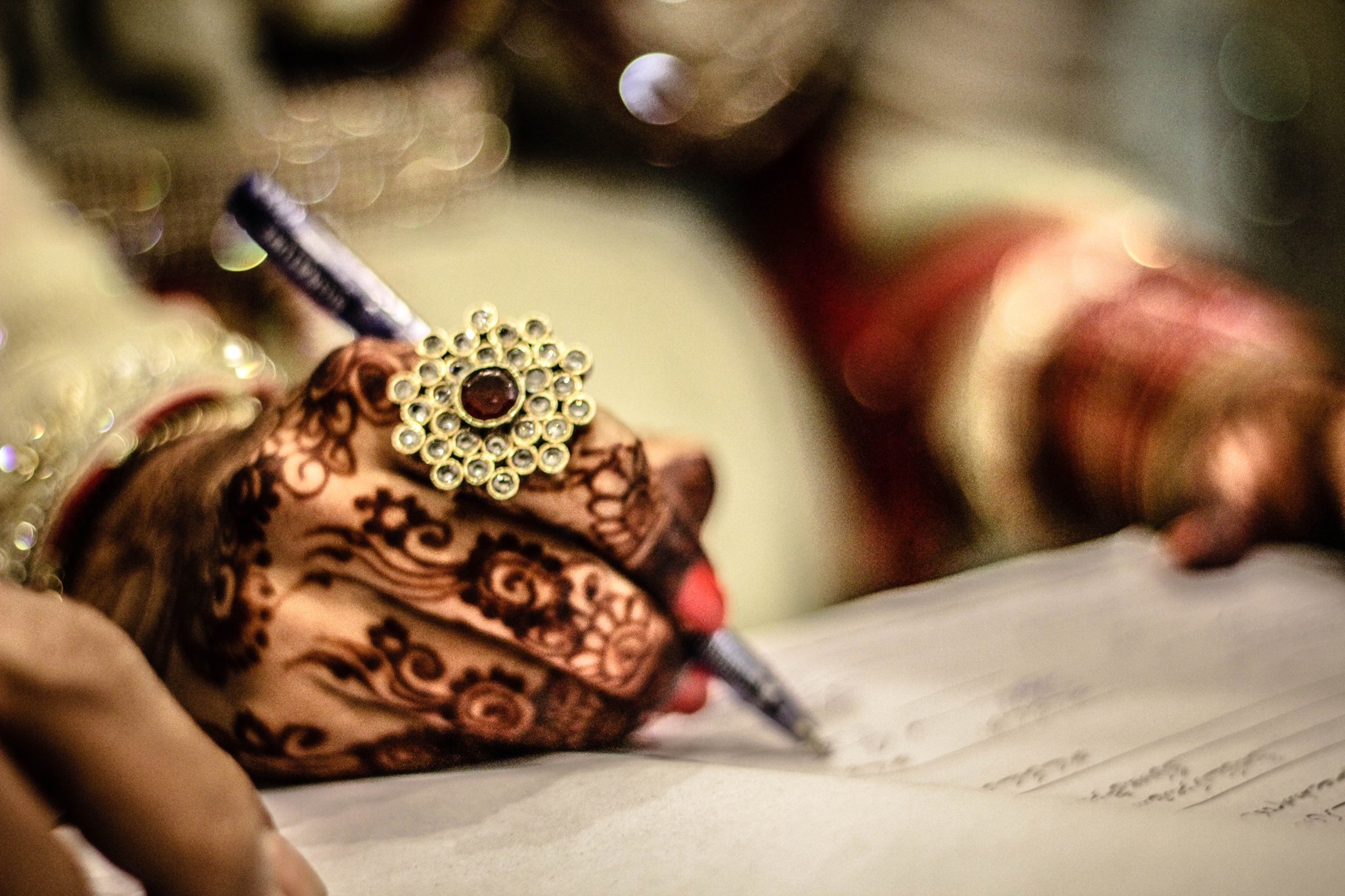 Close-up of a hand adorned with intricate henna designs and a jeweled ring, holding a pen and writing on a paper.