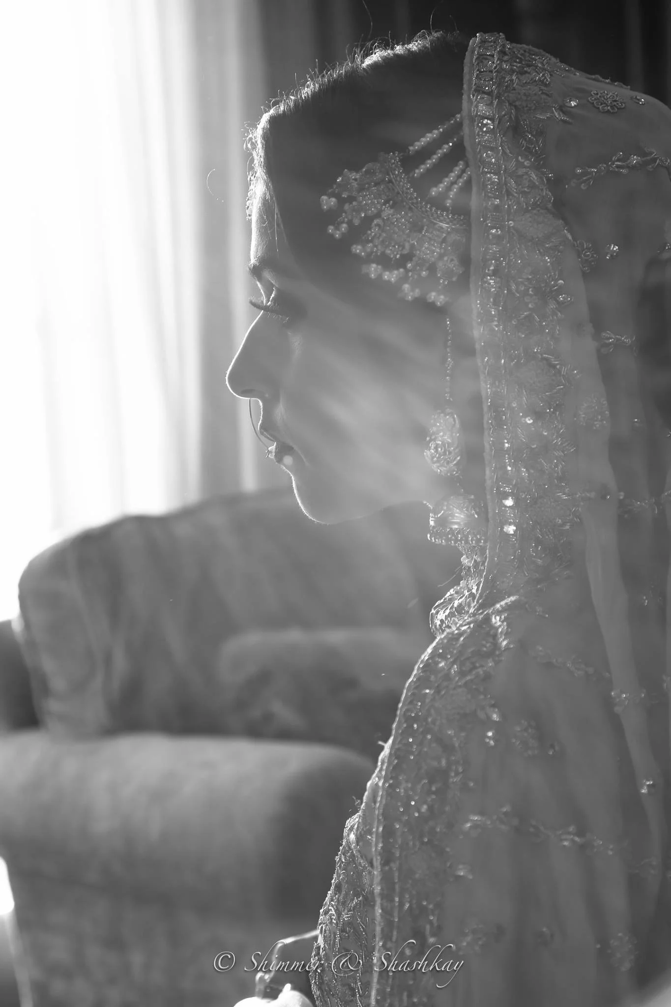 Profile of a woman wearing a traditional bridal outfit, adorned with intricate embroidery and jewelry, in a black and white setting, with soft light streaming through a curtain.