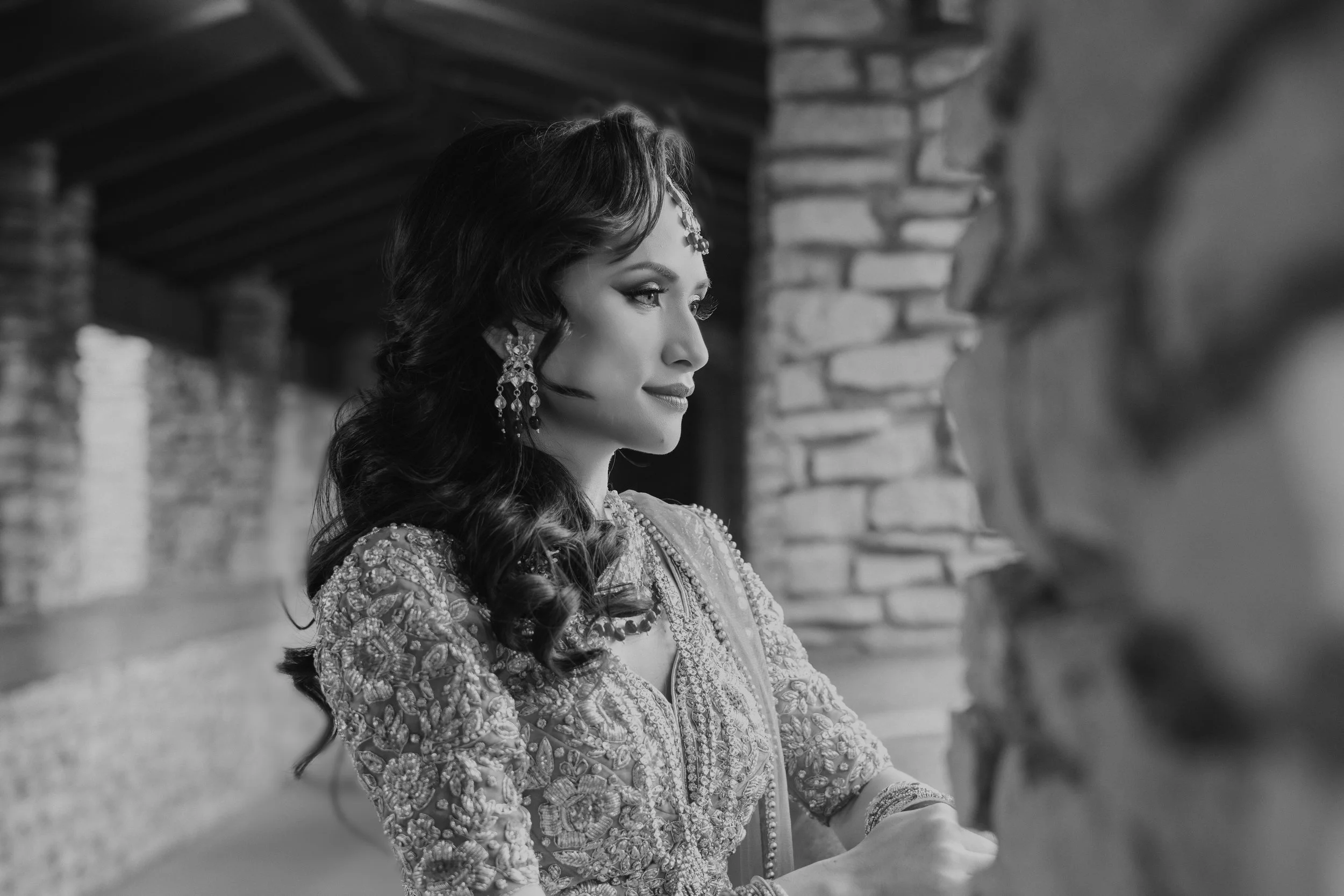 Black and white portrait of a woman in traditional attire with intricate embroidery, wearing large earrings and a headpiece, standing in a stone hallway.