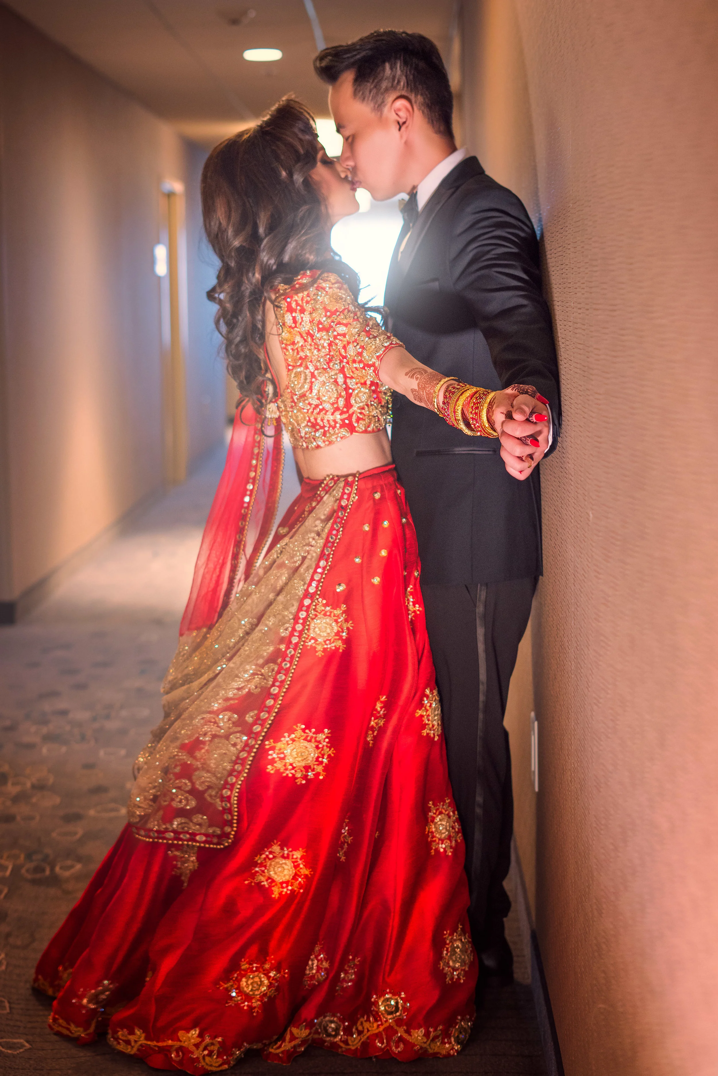 A couple kissing in a hallway, with the woman wearing a traditional red and gold outfit and the man in a black suit.