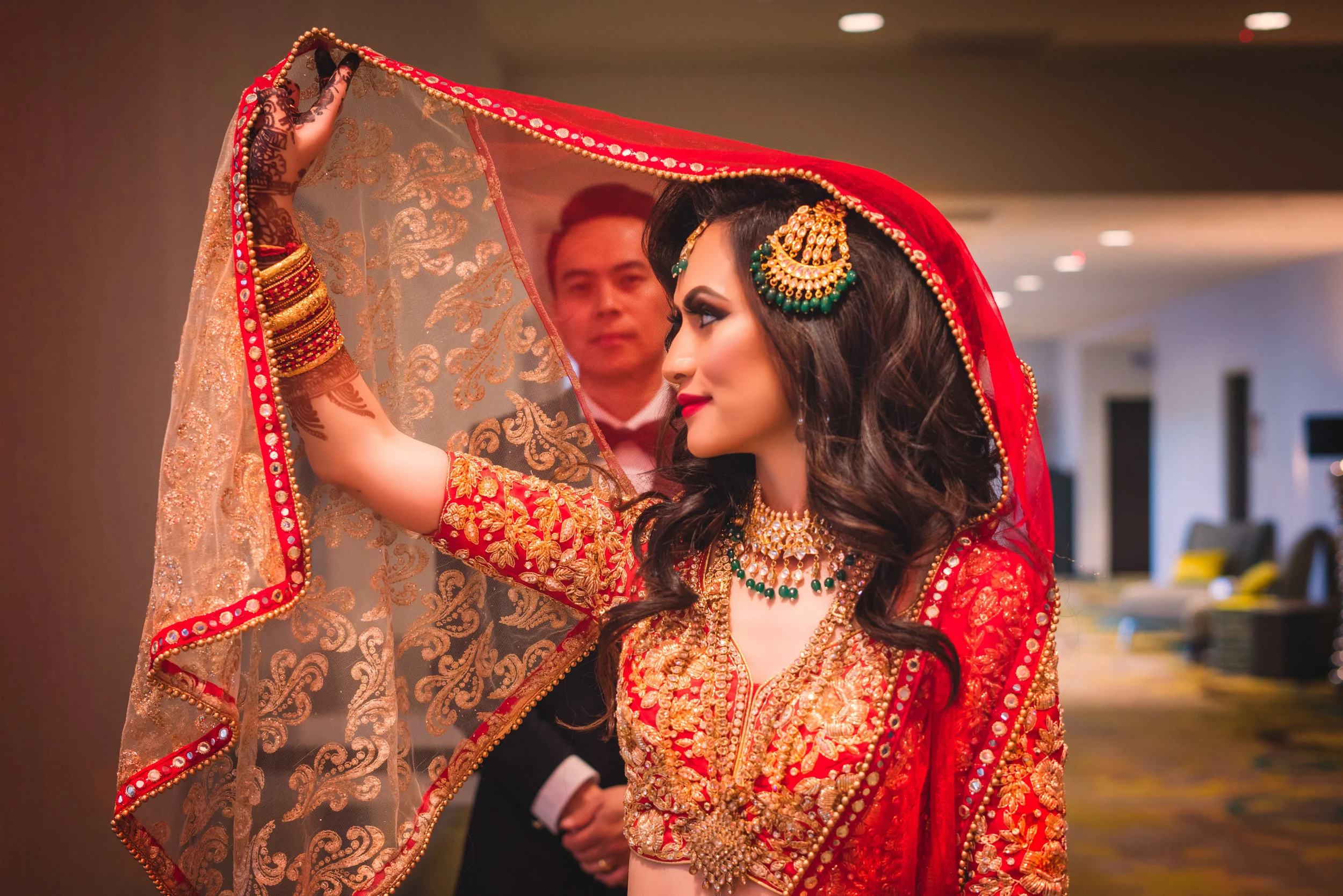 Bride wearing traditional red and gold South Asian attire, with intricate jewelry and henna, holding a decorated veil. A man stands in the background.