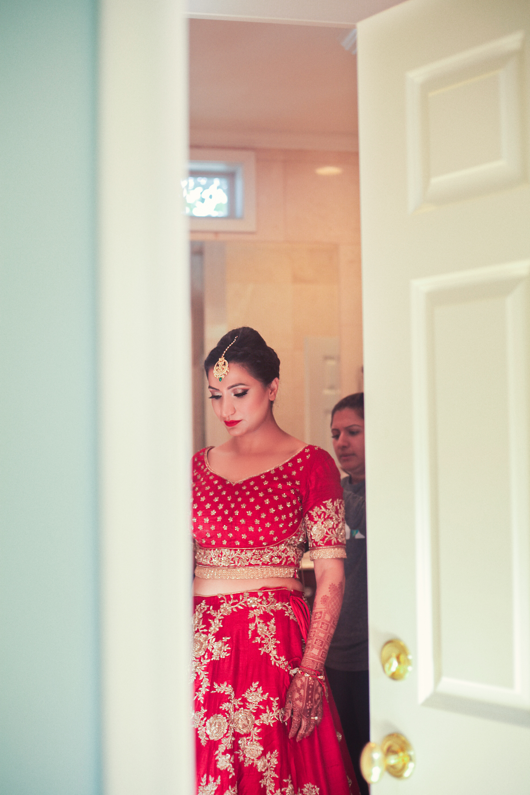 A woman in a red and gold traditional outfit with intricate embroidery, wearing a maang tikka and henna designs on her arms, stands partially visible through a slightly ajar door.